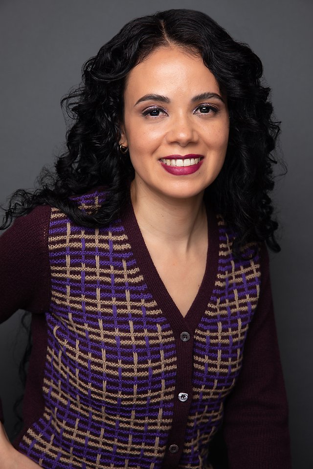 Smiling woman with curly black hair wearing a purple and beige patterned cardigan against a gray background