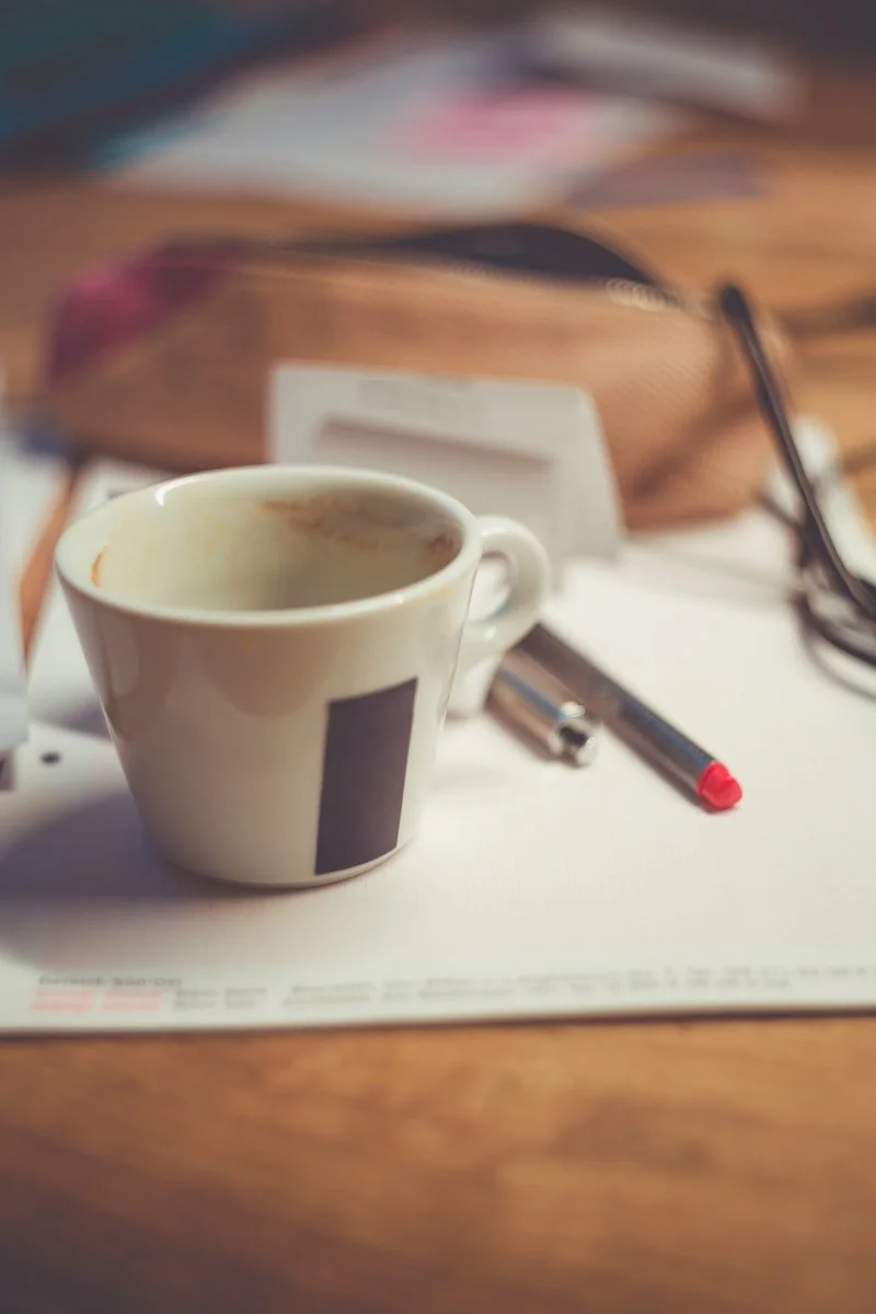 A partially used white coffee cup, a black pen with a red cap, and a pair of glasses on a desk with papers.