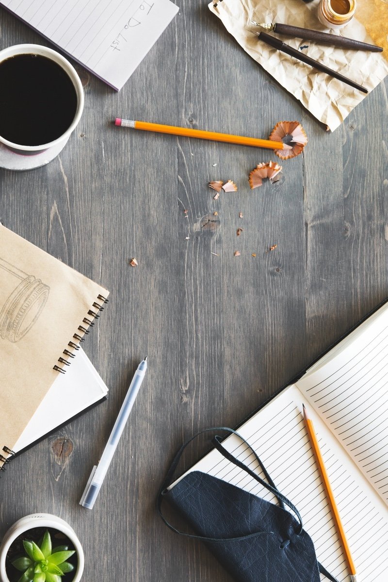 A cluttered wooden desk with a cup of black coffee, notebooks, pencils, a pen, and a small potted plant.
