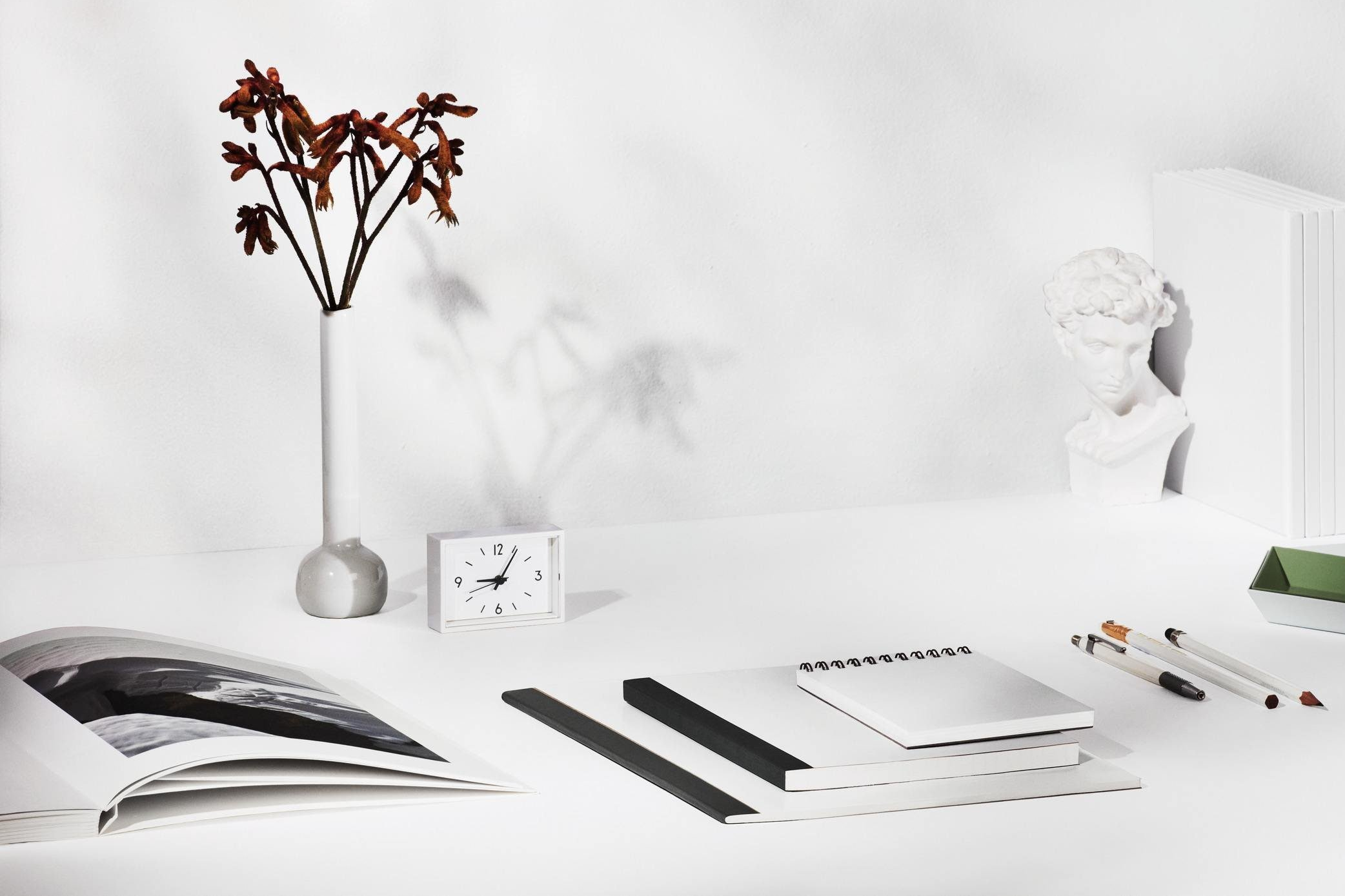 Minimalist white desk with dried flowers in a vase, books, clock, pencils, sketchbook, and classical bust sculpture