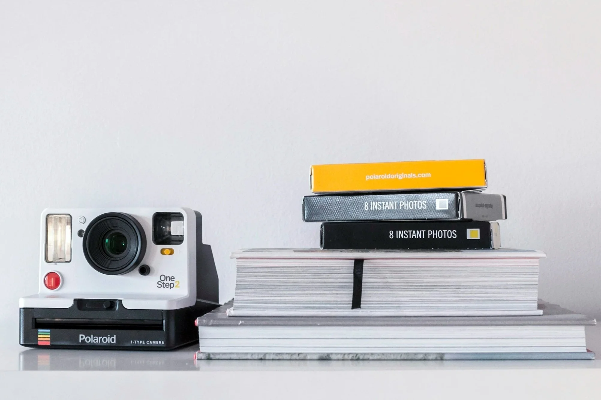 Polaroid camera and stacks of film and notebooks on a white surface against a plain white background.