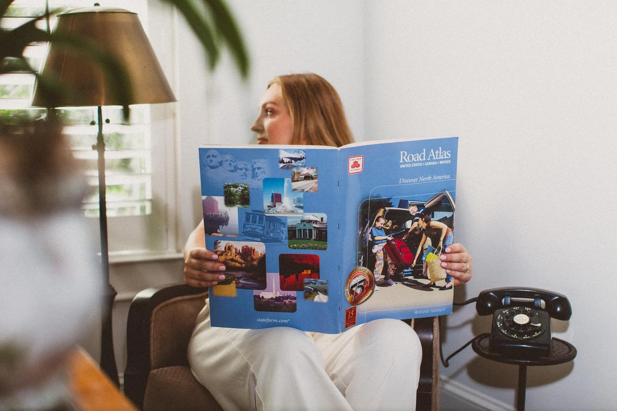 A woman with red hair sitting in a chair, reading a blue travel guide titled 'Road Atlas'. A vintage black rotary telephone is on a small side table next to her. A lamp and window with white shutters are also visible in the background.
