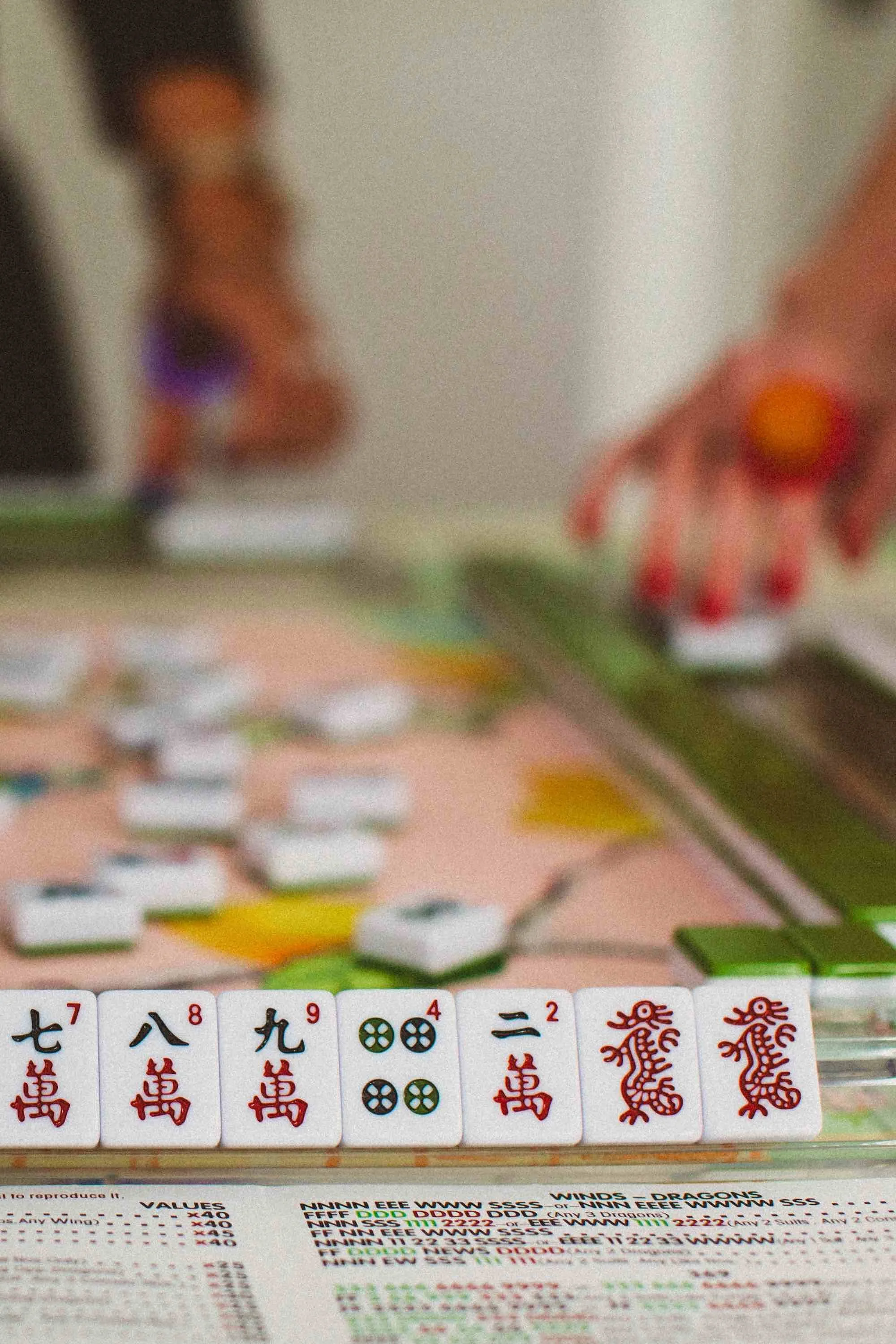 Close-up of mahjong tiles on a table with a person's hand reaching for a tile, blurred background.