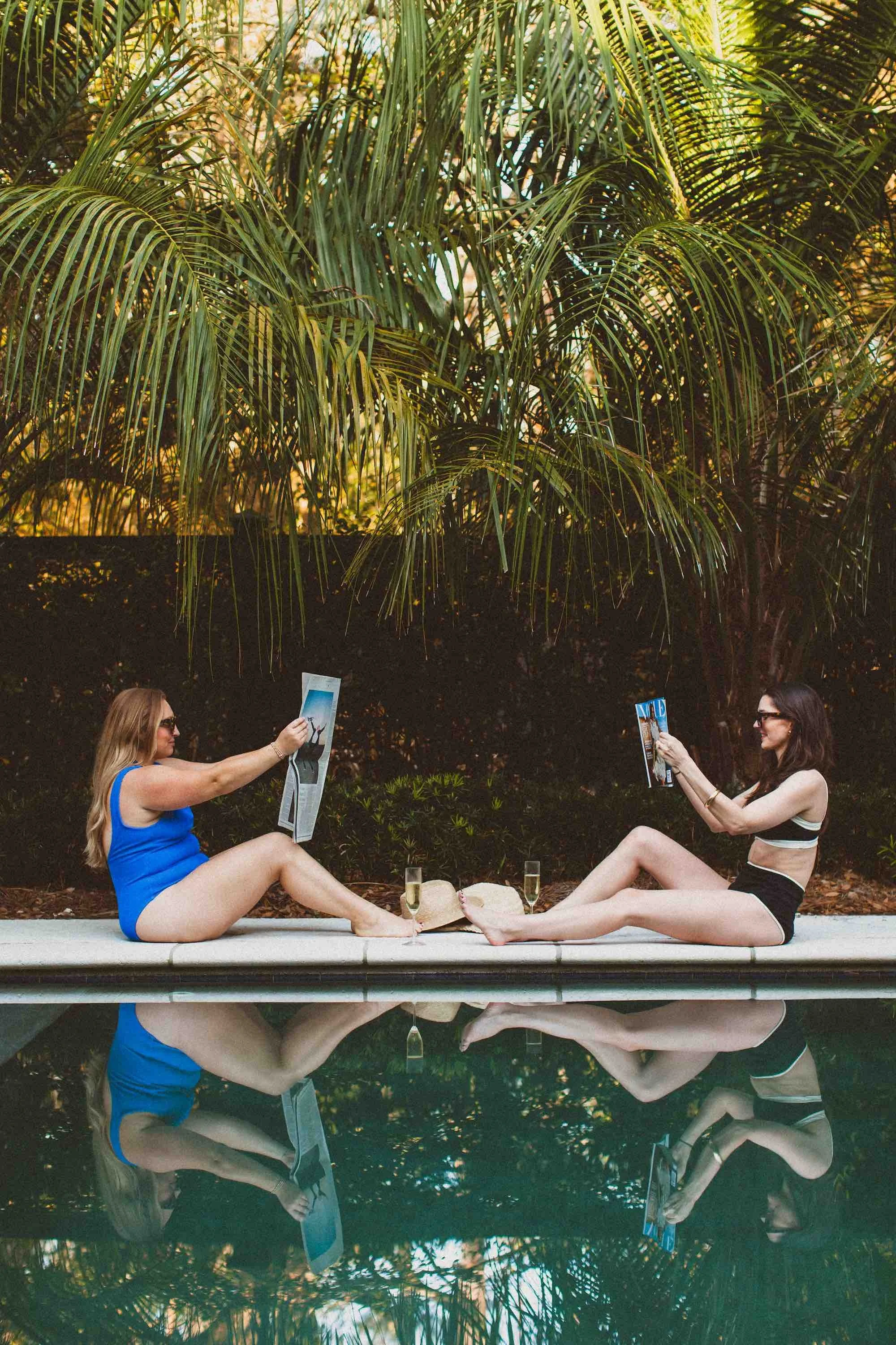 Two women in swimsuits sitting by a pool, reading magazines, with drinks and straw hats, surrounded by tropical plants.