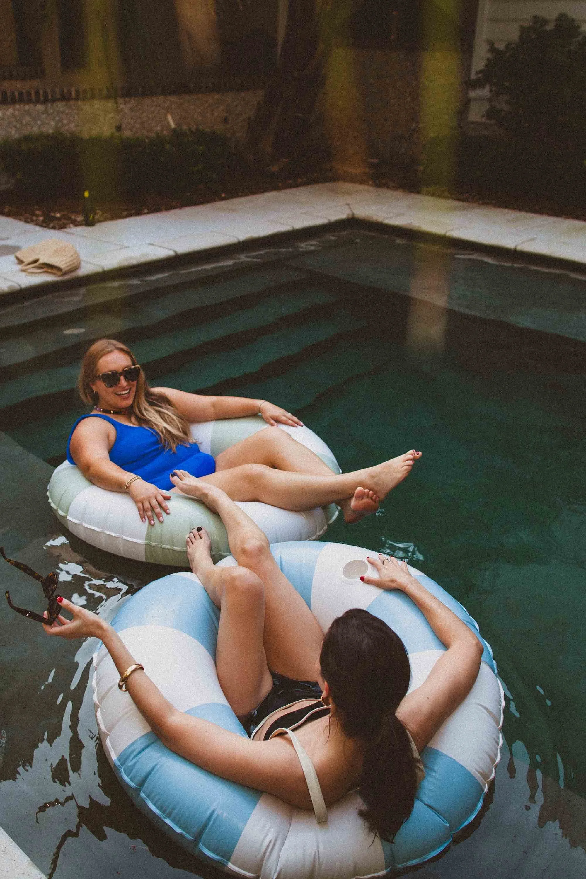 Two women relaxing on inflatable pool floats in a swimming pool, one woman wearing a blue swimsuit and sunglasses, smiling.