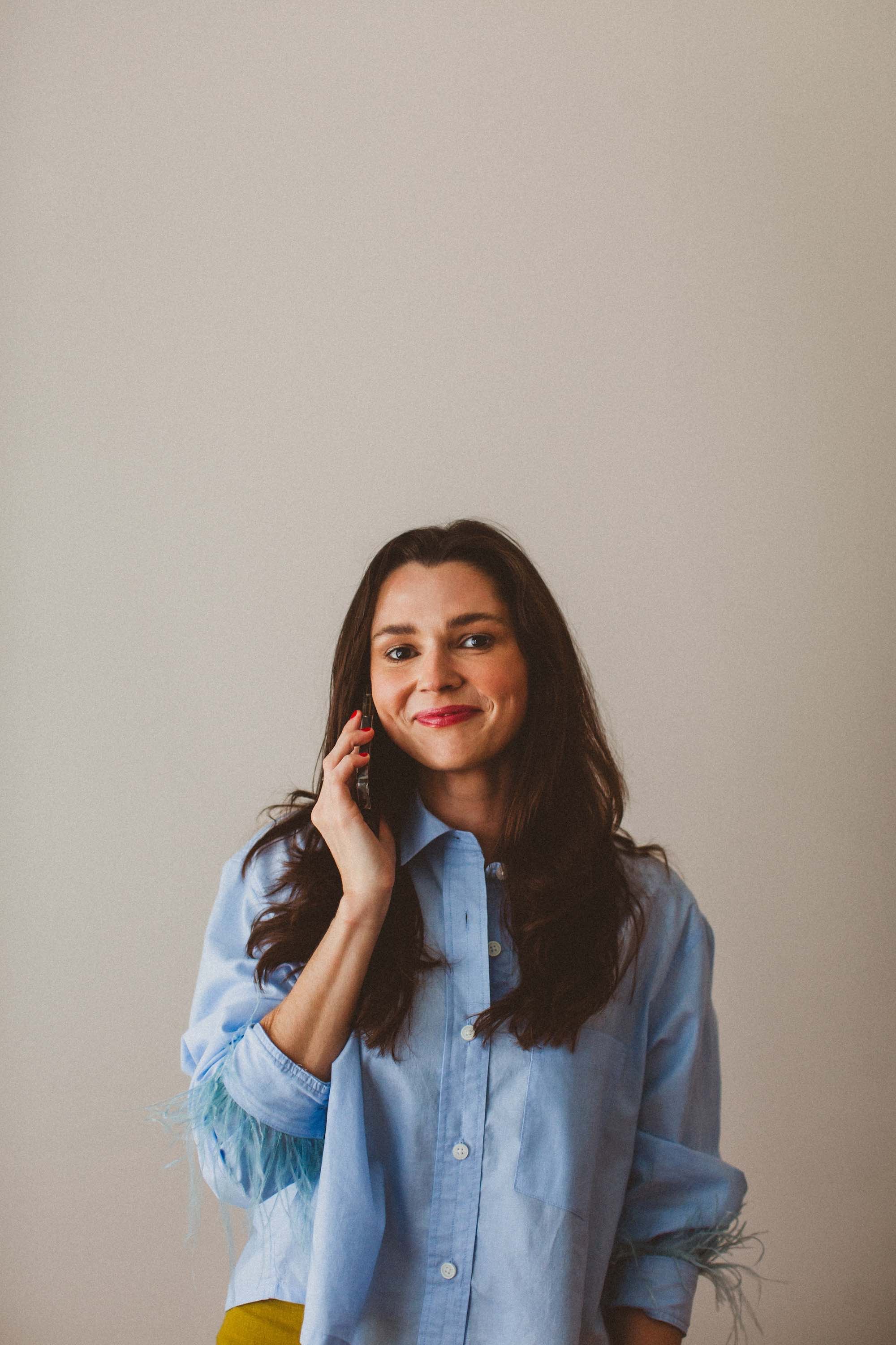 A woman with long brown hair, wearing a light blue button-up shirt, is smiling and talking on a mobile phone.