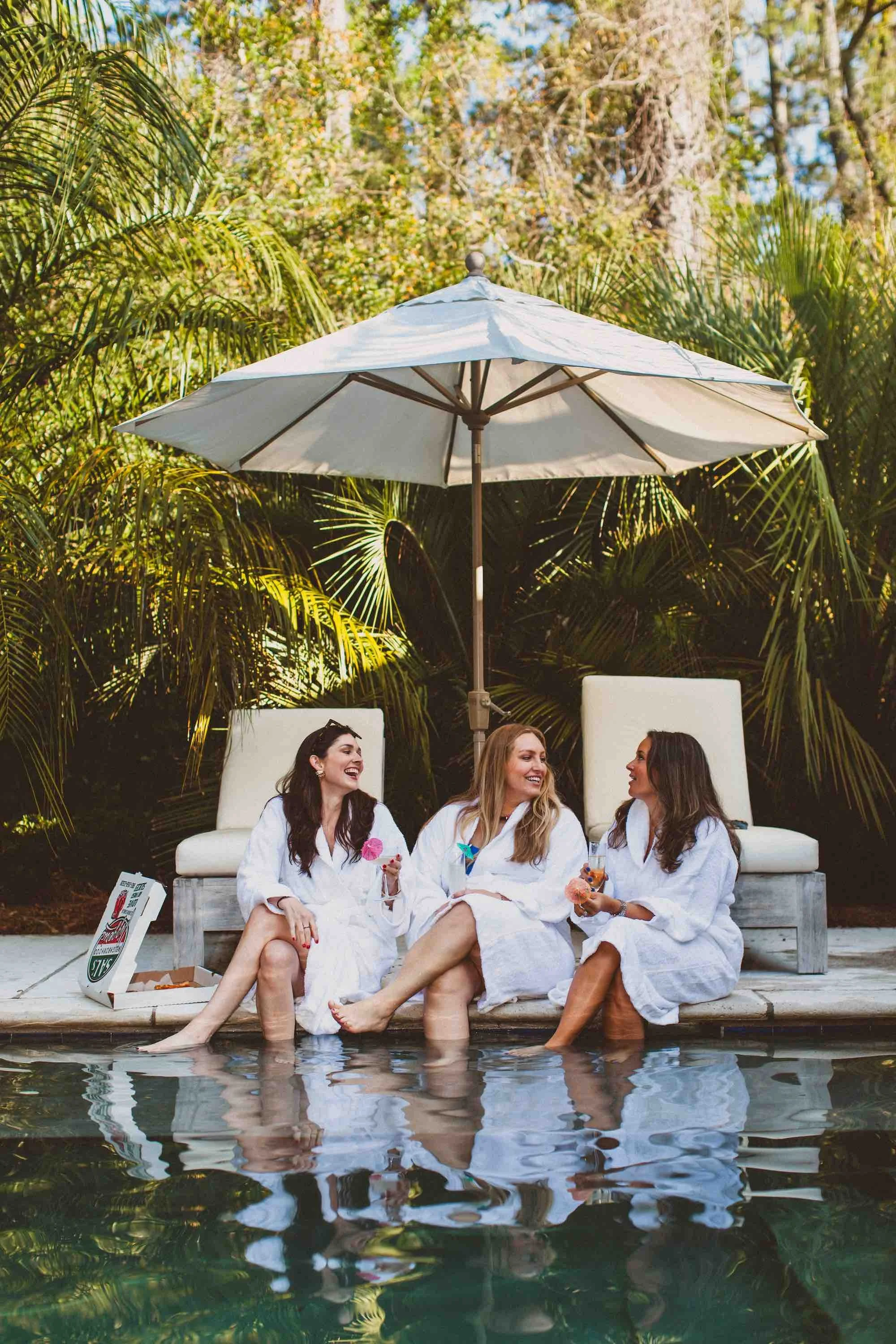 Three women in white robes sitting by a pool, laughing and chatting, with drinks in hand, under a large white umbrella surrounded by lush green bushes.