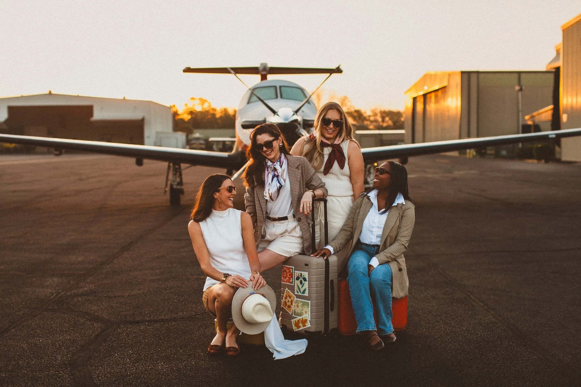 Group of five women with luggage smiling in front of small airplane at sunset.