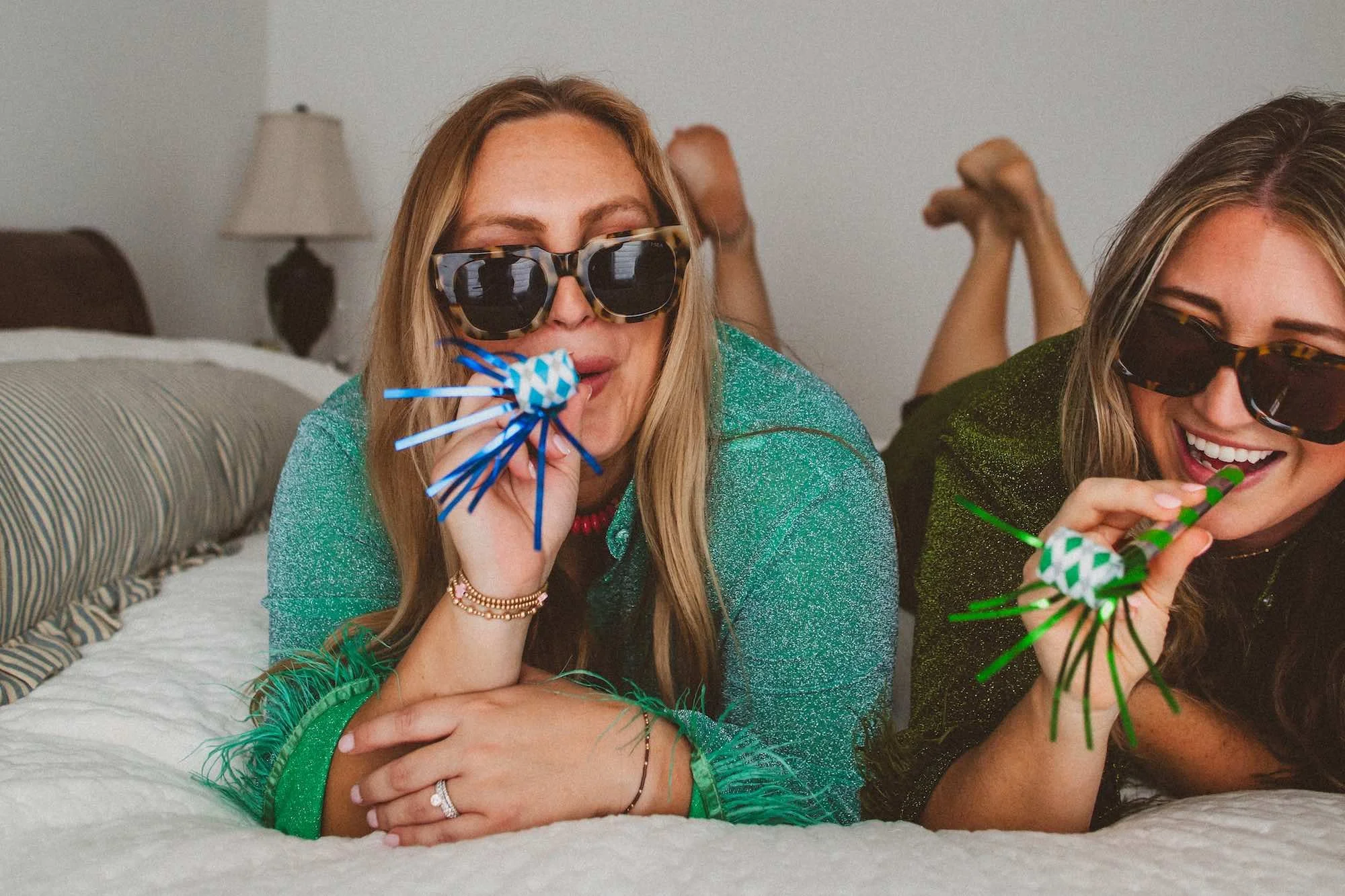 Two women wearing sunglasses and party outfits, lying on a bed, celebrating with party blowers and confetti.