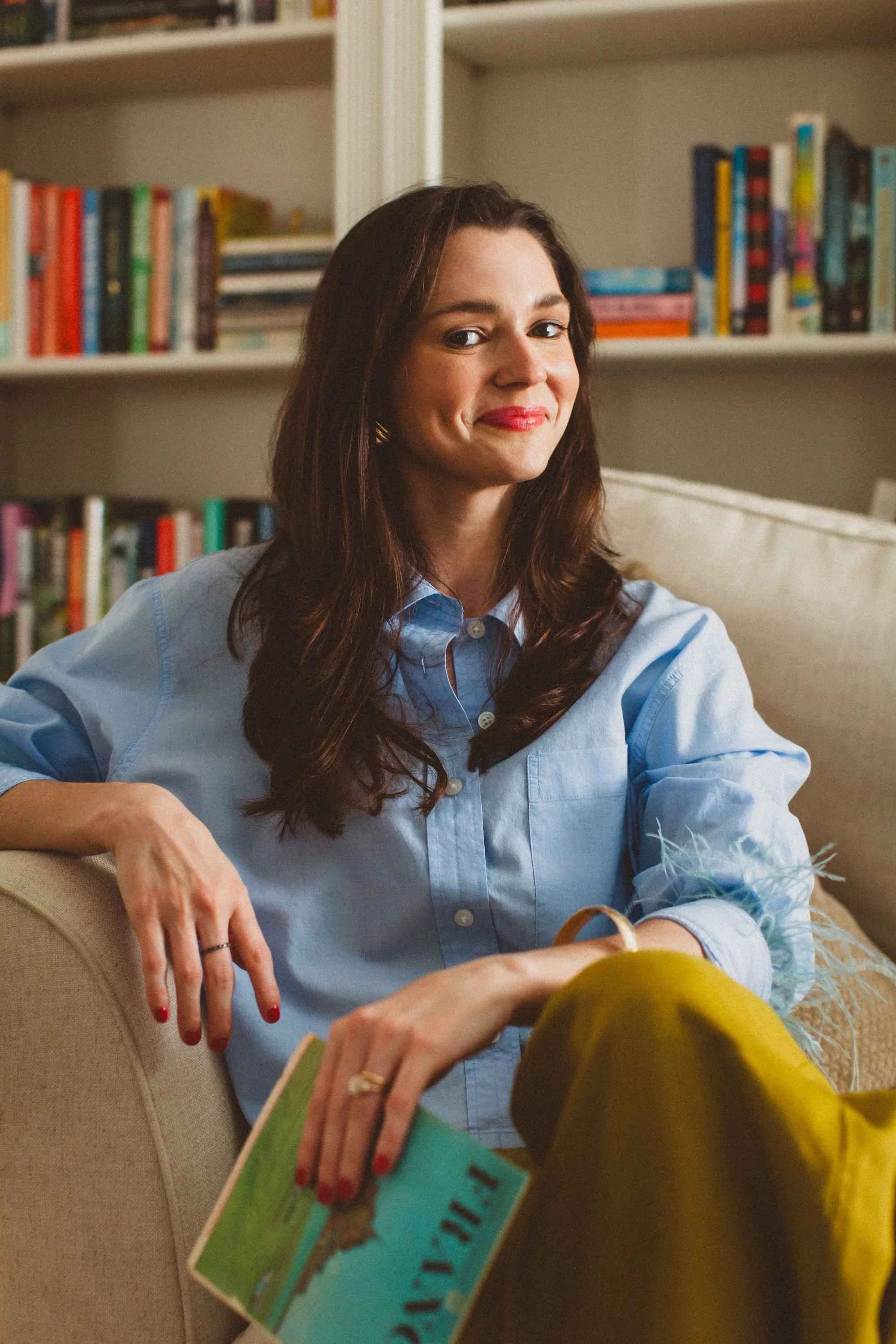 A woman sitting on a beige sofa, holding a book, with a background of bookshelves filled with colorful books.