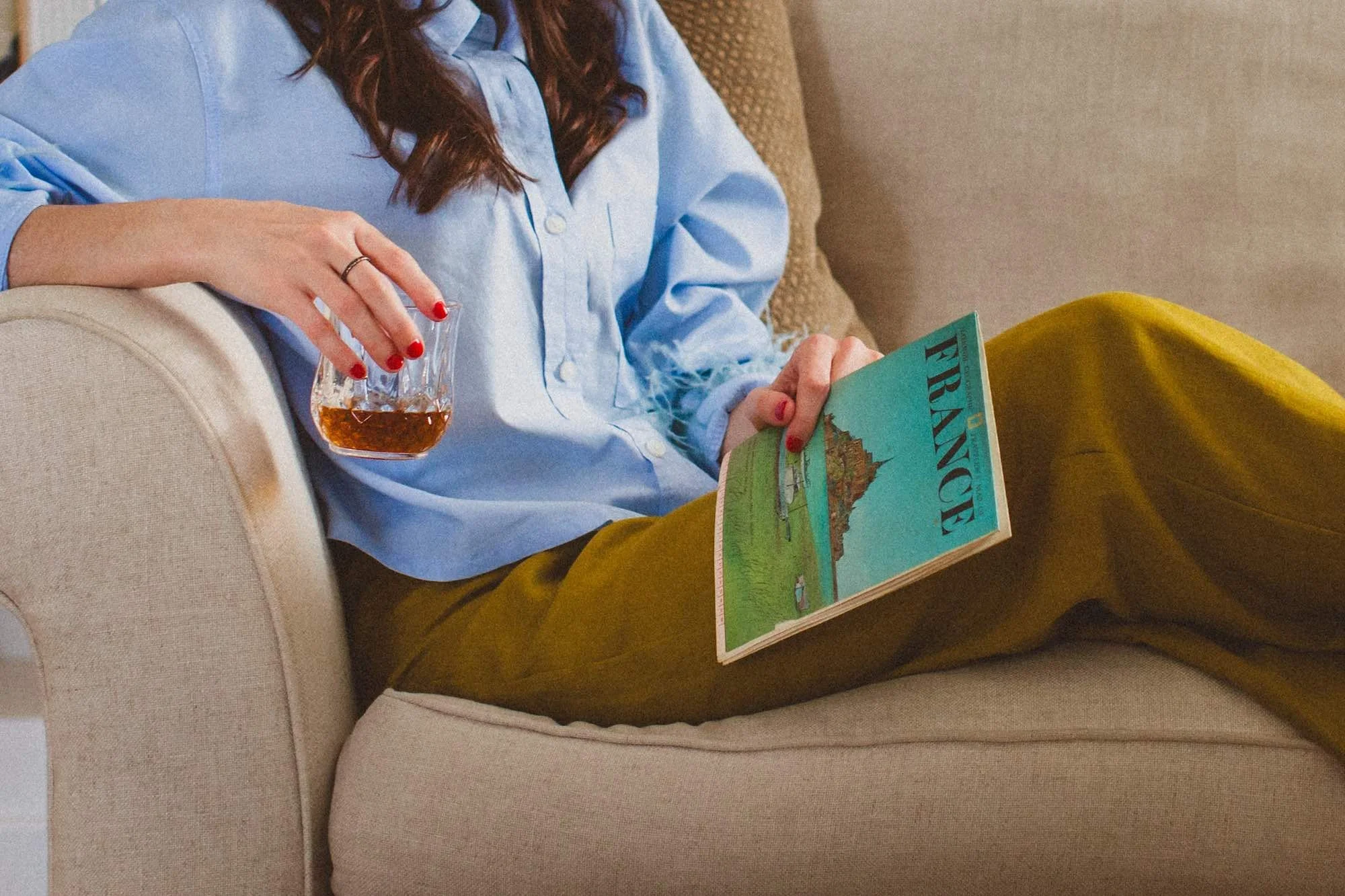 A woman with red hair, wearing a light blue shirt and mustard yellow pants, is sitting on a beige couch. She is holding a glass of amber-colored drink in her left hand and a magazine titled 'France' in her right hand.