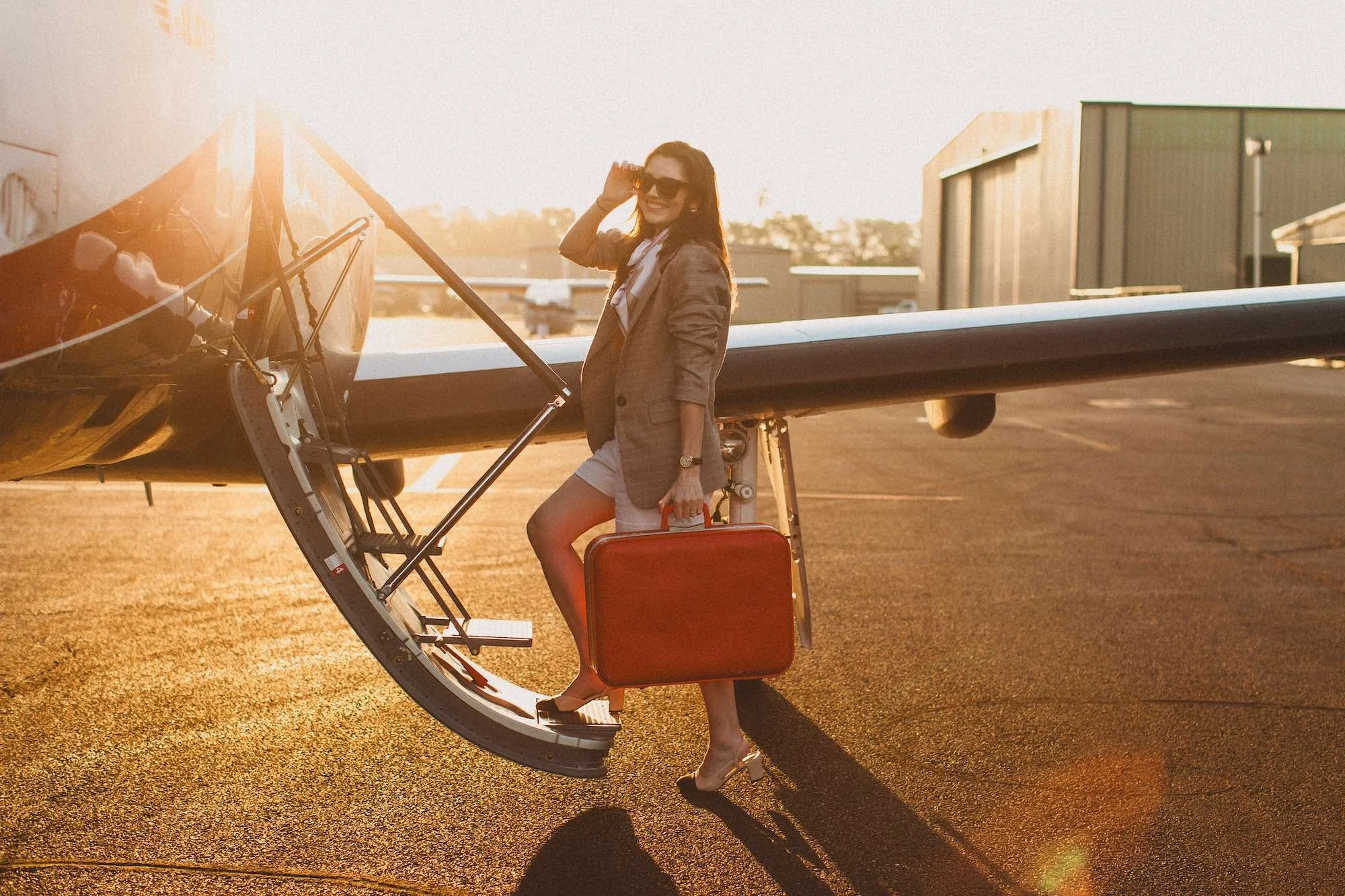 A woman wearing a blazer, shorts, high heels, and sunglasses stands on the staircase of a small private airplane, holding a red suitcase in one hand, as the sun sets in the background.