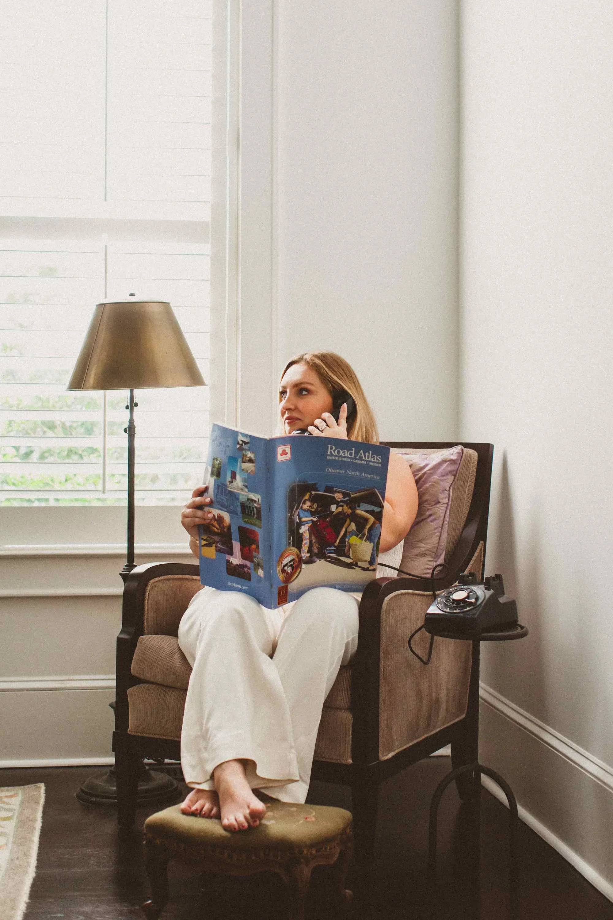 A woman with long blonde hair sitting on a beige armchair, talking on a phone while holding a travel guidebook titled 'Road Atlas'. She is in a bright room with large windows, and there is a small side table with a rotary phone next to her. She is ba
