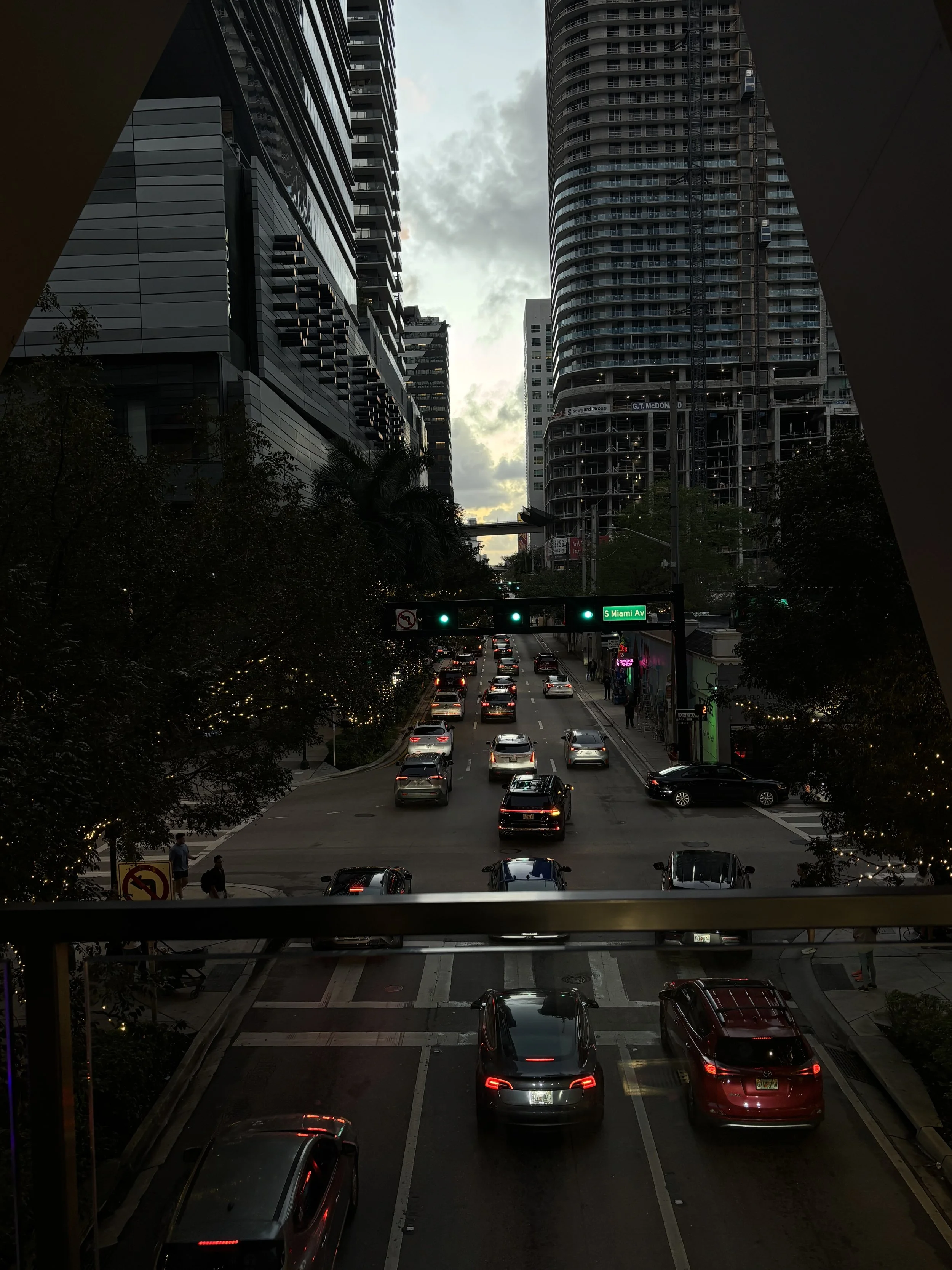 City street scene at dusk with traffic, tall buildings, trees, and a sign indicating South Miami Avenue.