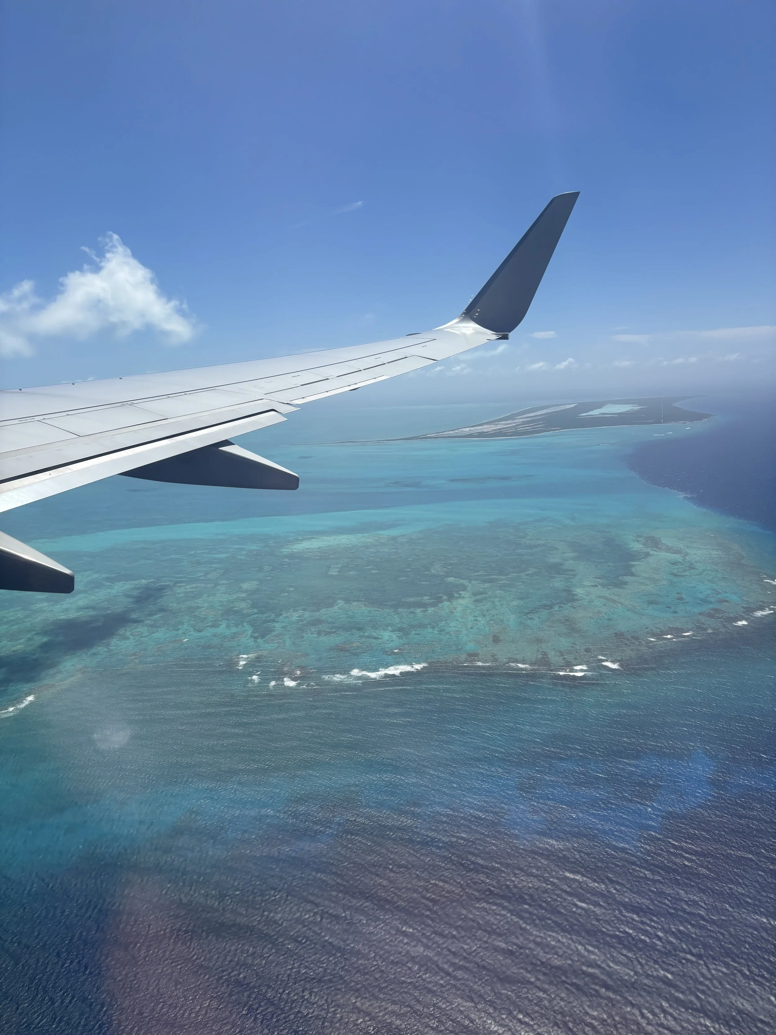 View from an airplane window showing part of the wing and a tropical overwater island surrounded by turquoise water.