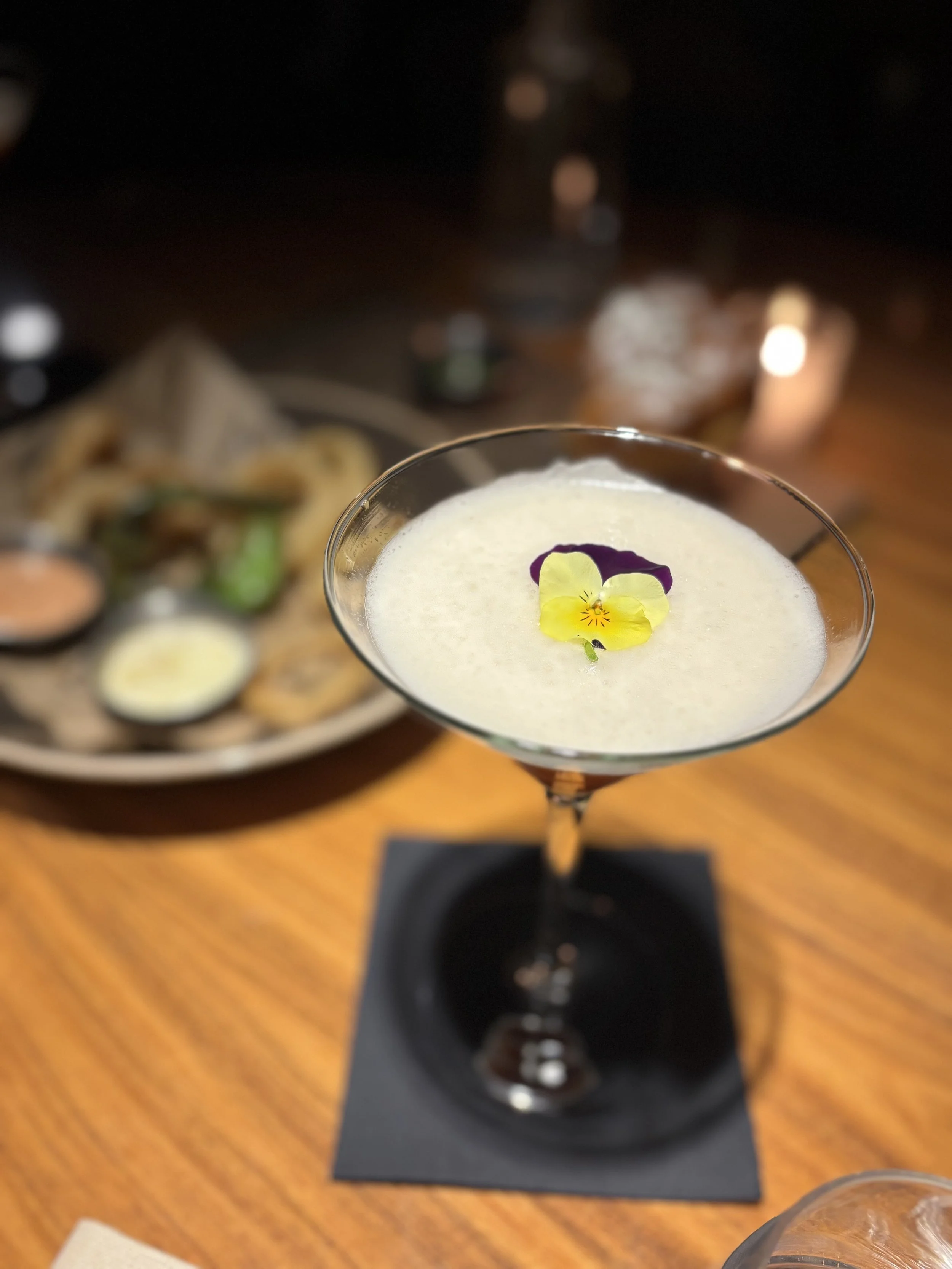 A cocktail in a martini glass garnished with edible flowers, placed on a black napkin on a wooden table with a blurred plate of food in the background.