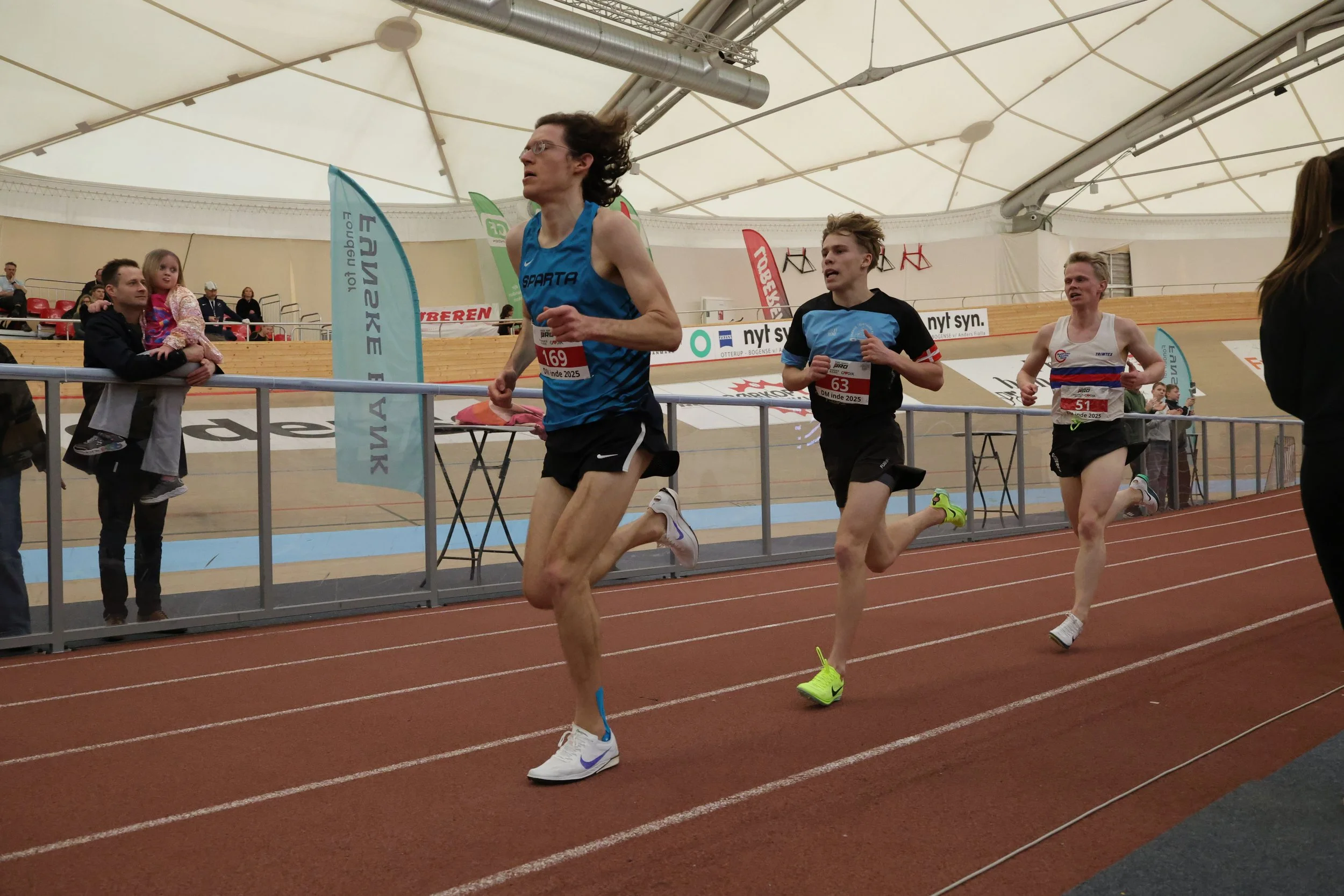 Three male athletes running on an indoor track during a race, with spectators watching from the side.