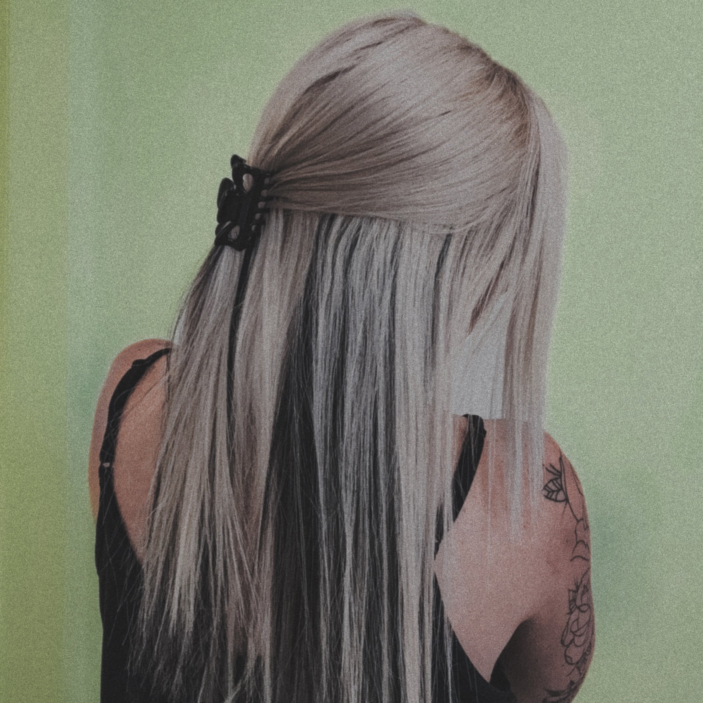 Back of a woman with long blonde hair held with a black hair clip, wearing a black sleeveless top, and showcasing floral tattoo on her upper arm against a green background.