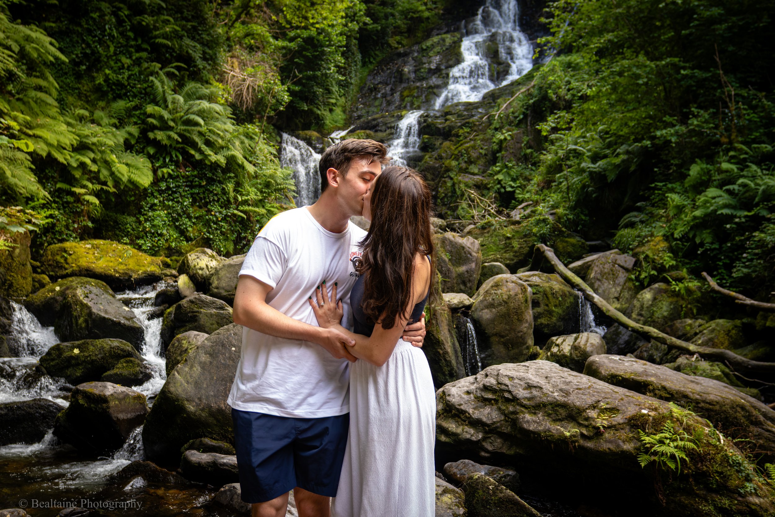 A couple kissing near a waterfall in a lush green forest, standing on rocks surrounded by flowing water.