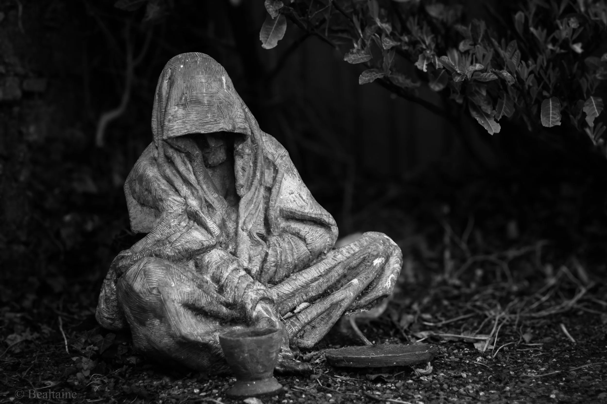 A black-and-white photo of a statue of a seated figure with a hooded head, with hands resting on its lap, placed outdoors among bushes and plants.
