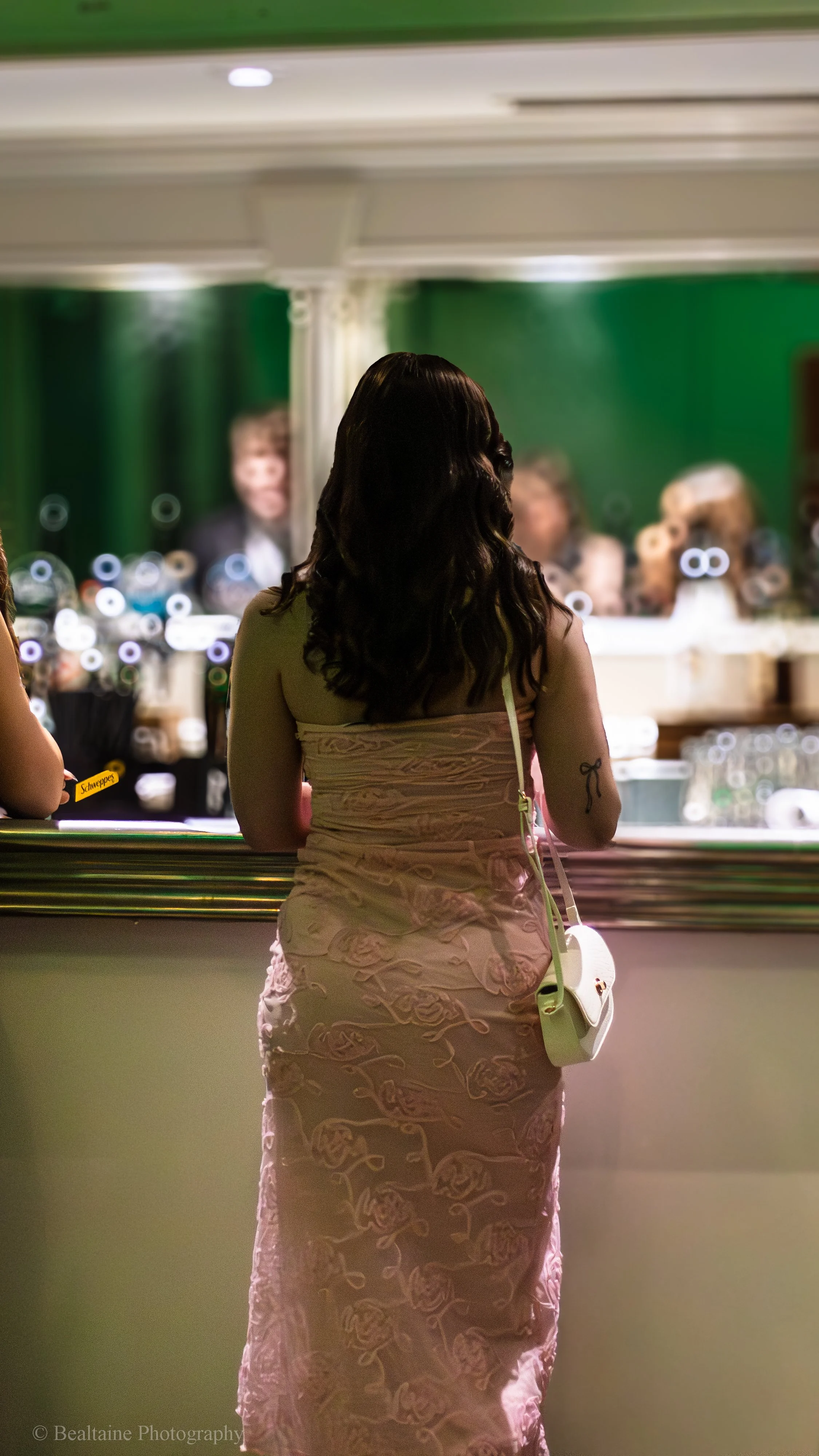 A woman with dark, wavy hair wearing a pink, embroidered dress stands at a bar or counter, looking towards a mirror or window with people visible in the background.