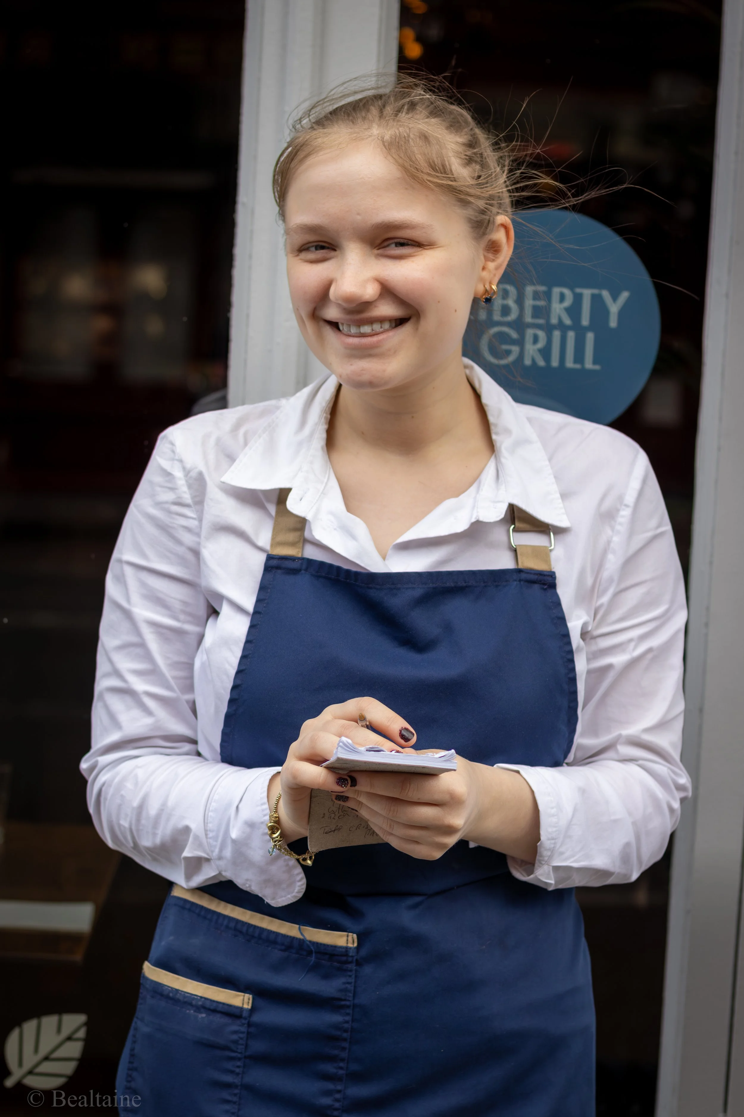 A smiling young woman with blonde hair, wearing a white shirt and a blue apron, stands in front of a window that has a sign reading 'Liberty Grill.' She is holding a notepad and a pen, with her hands clasped in front of her.