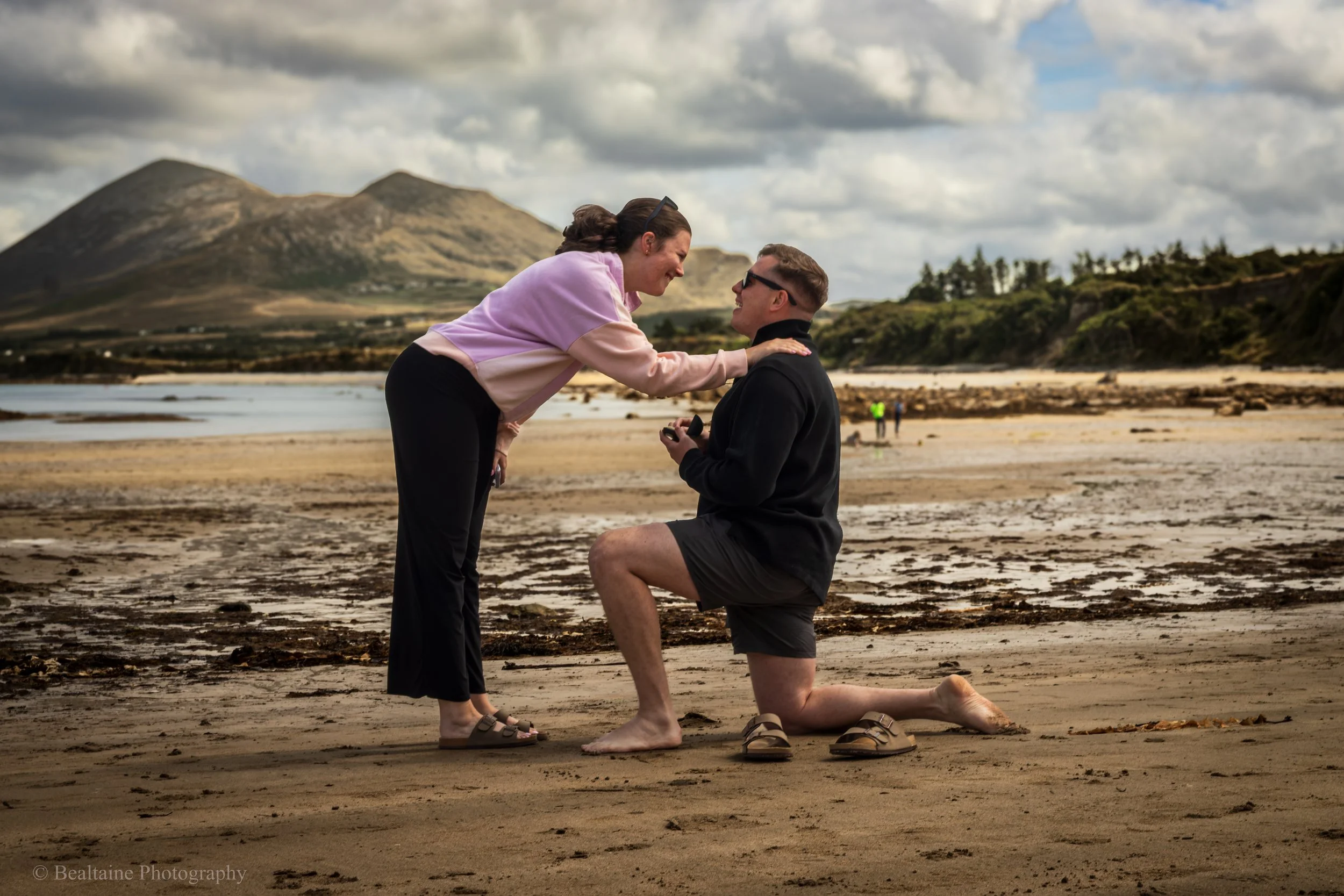 A woman is proposing marriage to a man on a beach, with mountains and cloudy skies in the background. The man is kneeling on one knee holding a ring box, while the woman leans forward holding his shoulders.