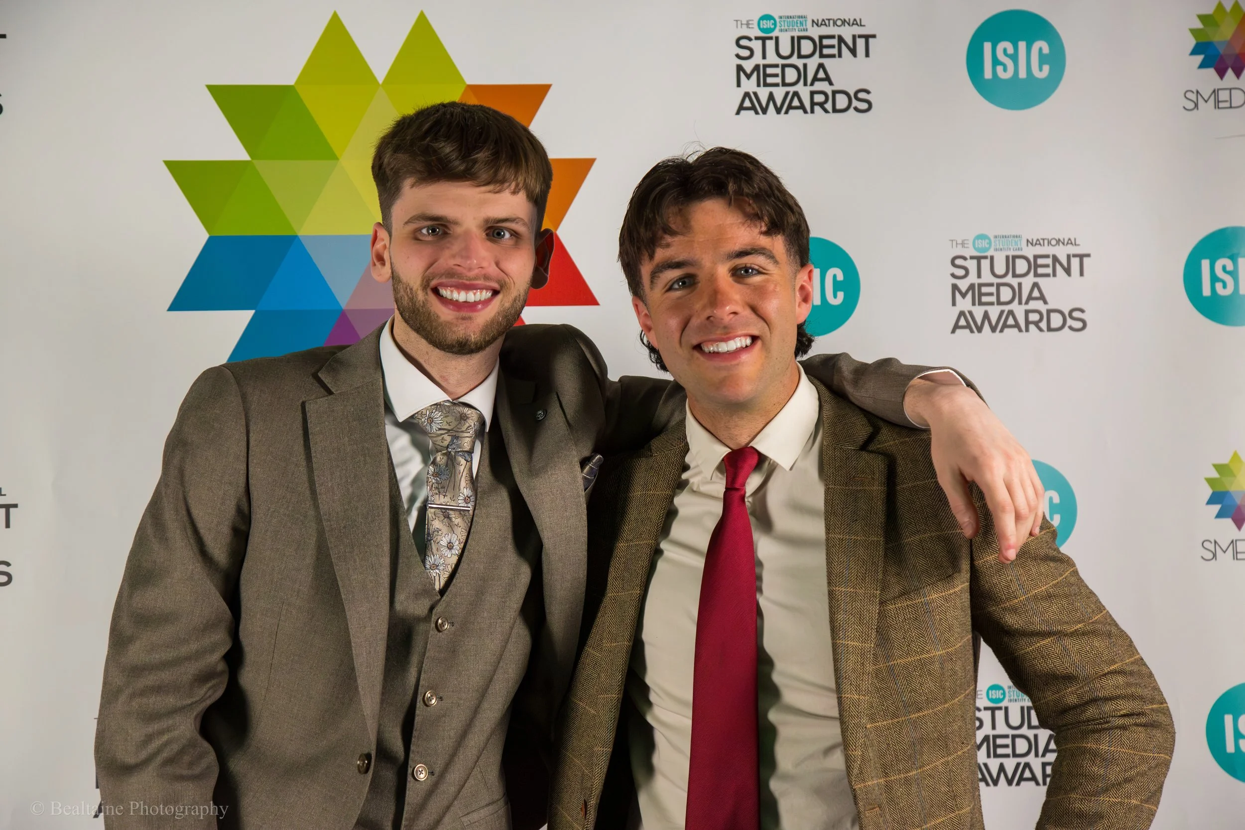 Two young men in suits smiling and posing together at the ISIC Student Media Awards event. The background features a colorful geometric pattern and the event's logo.