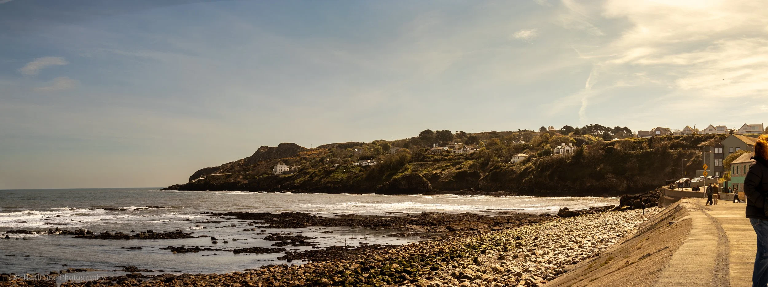 A coastal scene with a rocky beach, ocean waves, and a hillside with houses. A paved walkway runs along the shoreline with people walking and standing.