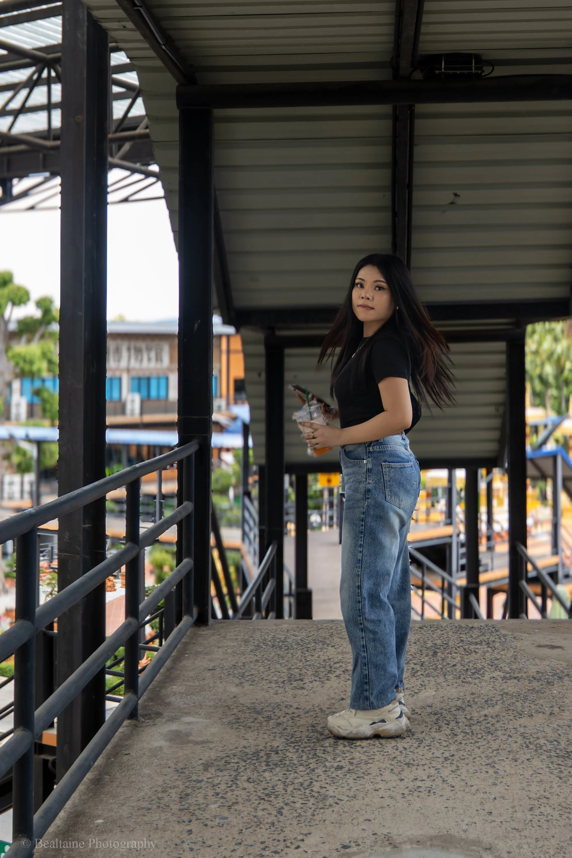 A young woman with long dark hair wearing a black shirt and high-waisted jeans standing on a balcony with metal railings, holding a drink and a phone, with a background of buildings and trees.