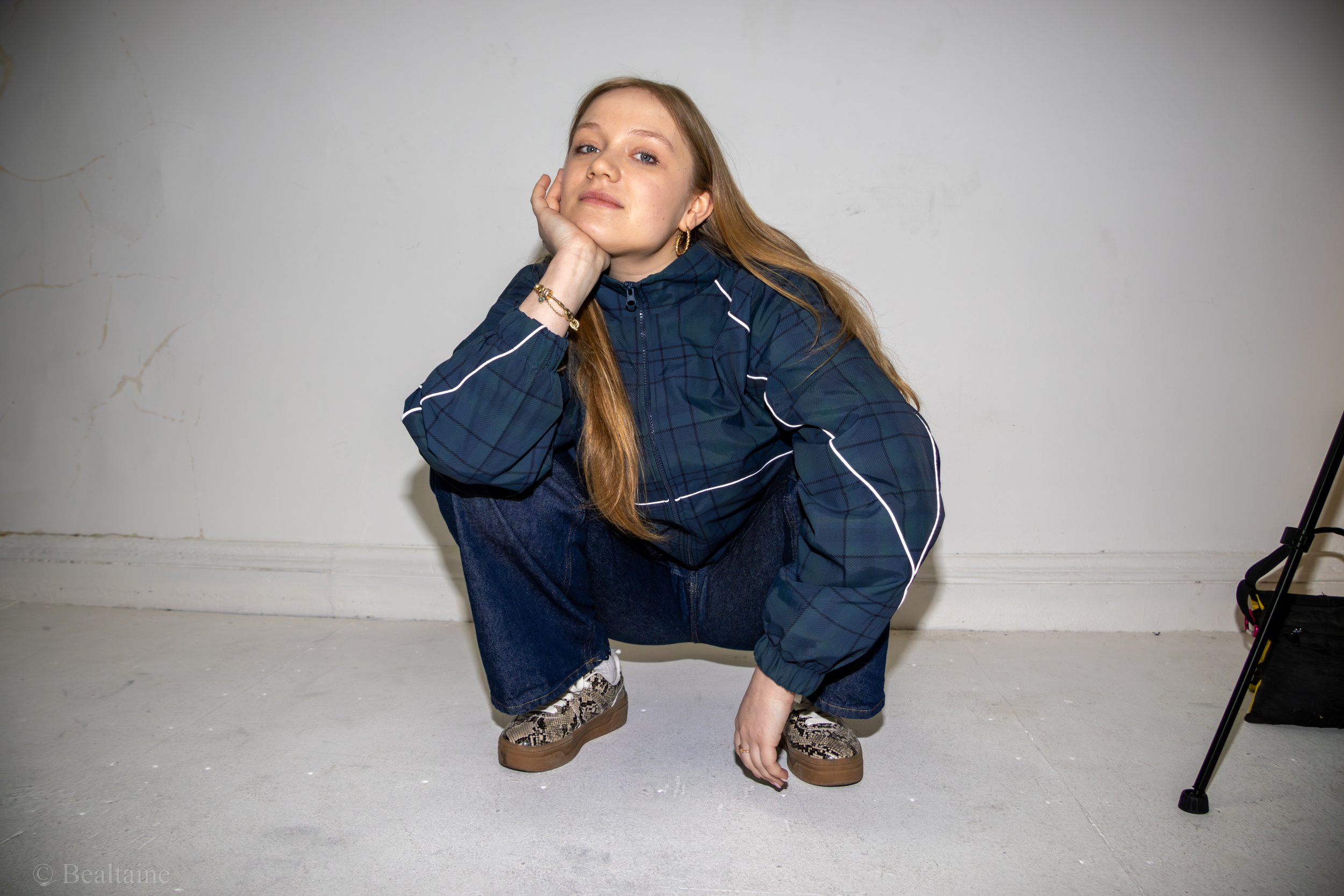 Young woman with red hair crouching, resting her face on her hand, wearing a blue track jacket with white stripes, blue jeans, snake print shoes, and gold jewelry, against a plain white wall.