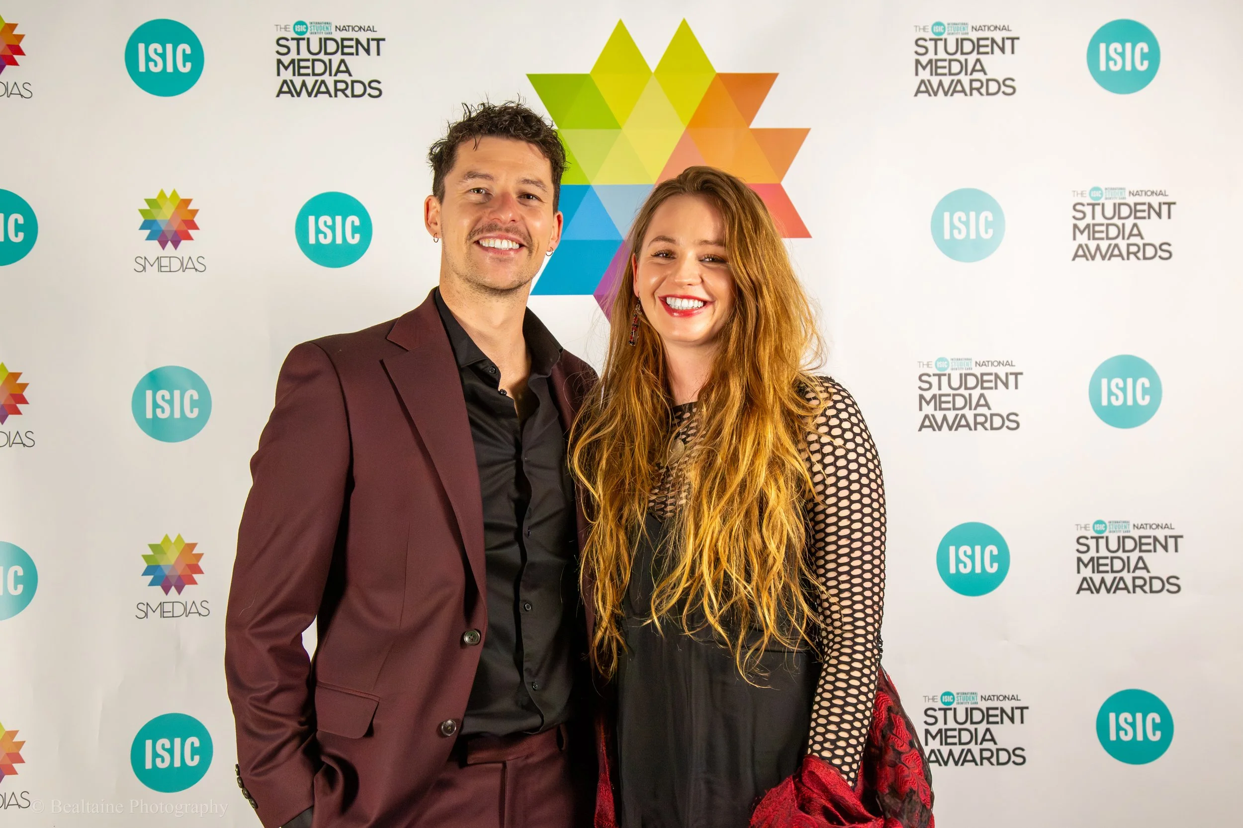 A young man and woman smiling at the camera at an awards event with a colorful geometric backdrop featuring logos for ISIC, SMIDAS, and the Student Media Awards.