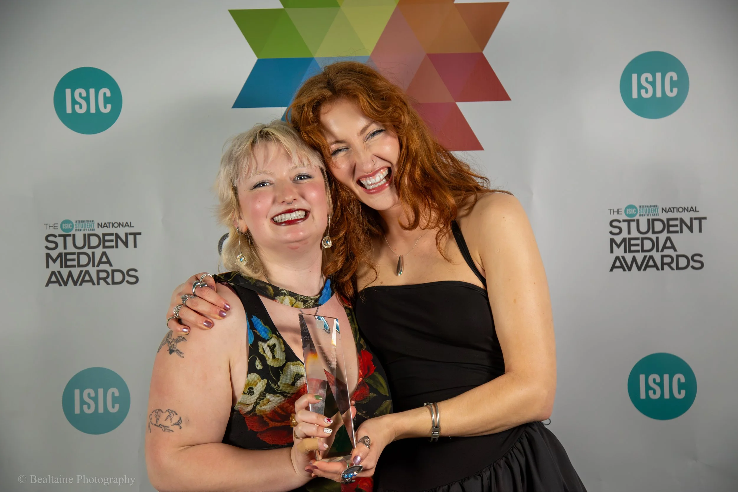 Two women smiling at the camera, one holding a crystal award, at the ISAIC Student Media Awards event with a backdrop featuring logos and colorful geometric shapes.