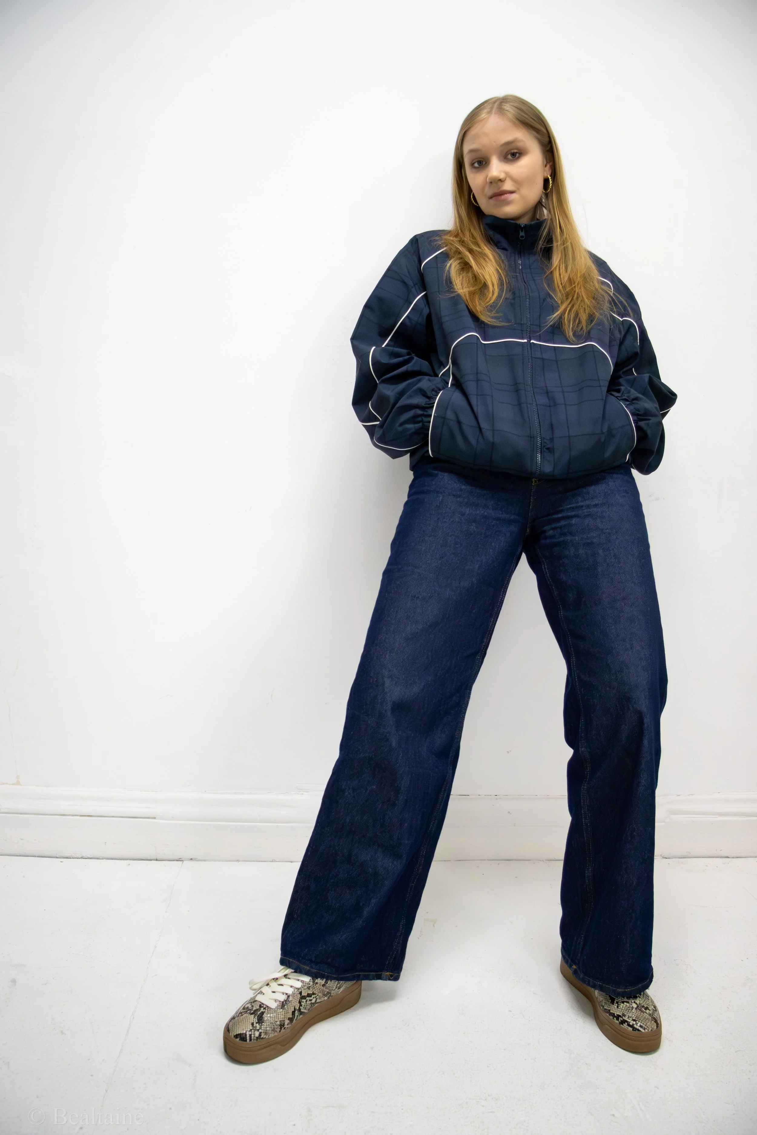 A young woman with long blonde hair wearing a navy athletic jacket, wide-leg jeans, and snake print sneakers stands against a plain white wall.