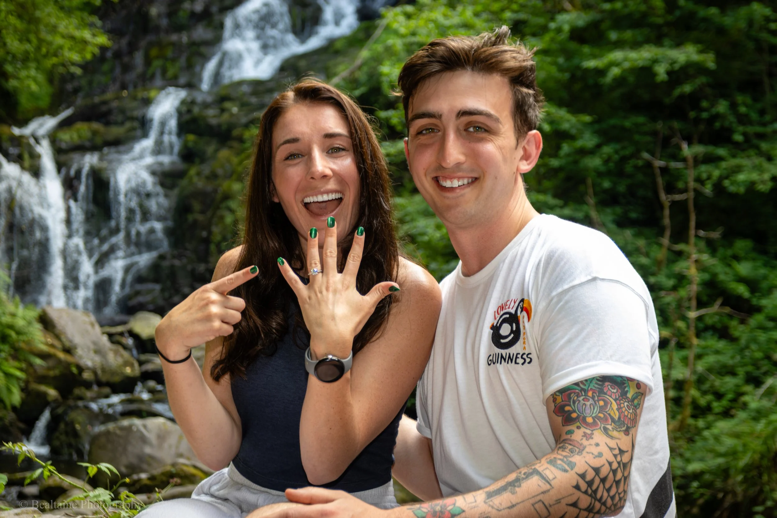 A smiling young man and woman taking a selfie in front of a waterfall surrounded by green foliage. The woman has long brown hair, visible rings and painted nails, and the man has short brown hair, tattoos on his arms and is wearing a white T-shirt wi
