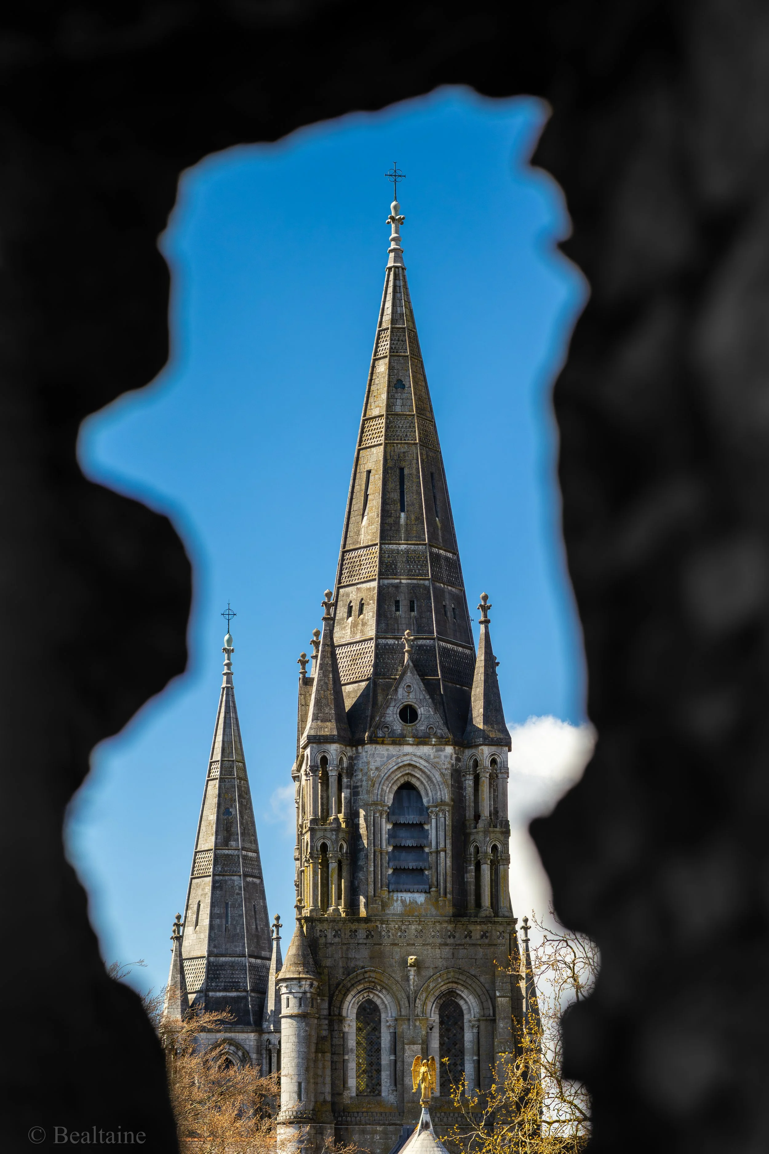 View of a gothic-style church tower with spires, seen through a hole in a dark structure, with a bright blue sky in the background.