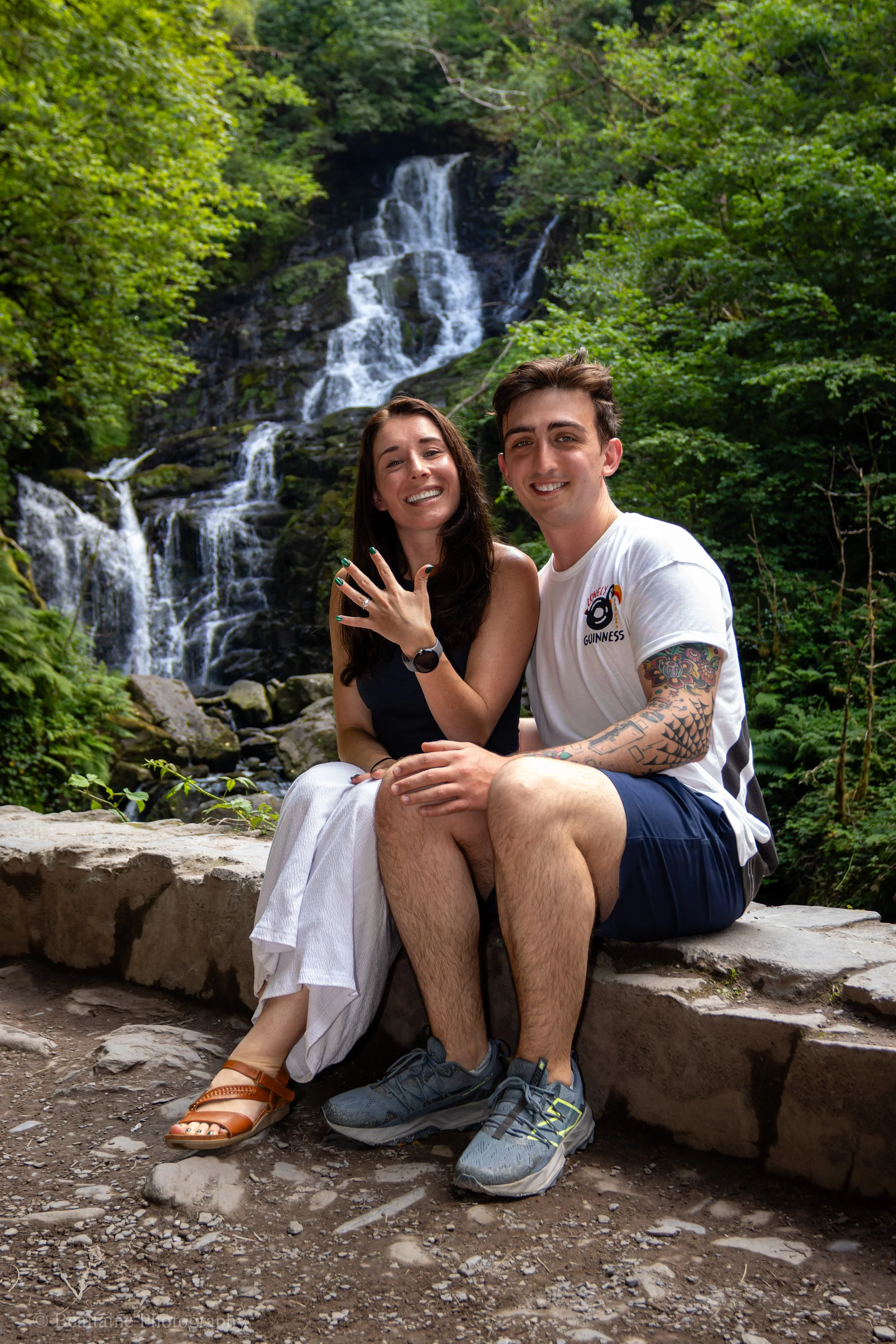 A smiling young couple sitting on rocks by a waterfall in a lush green forest.