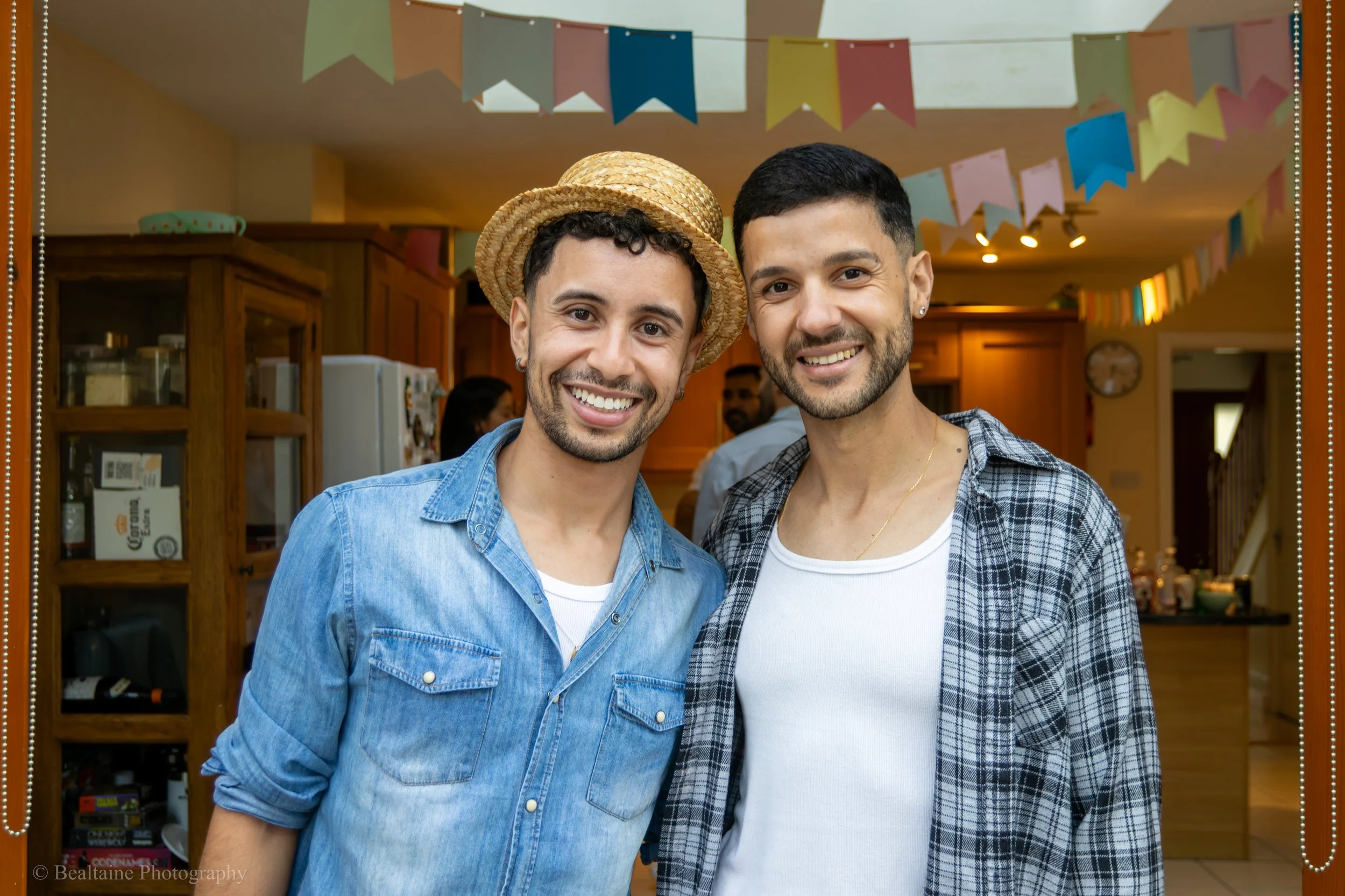 Two smiling men, one wearing a straw hat and a denim shirt, and the other in a plaid shirt over a white tee, are standing close together in a party setting decorated with colorful paper flags.