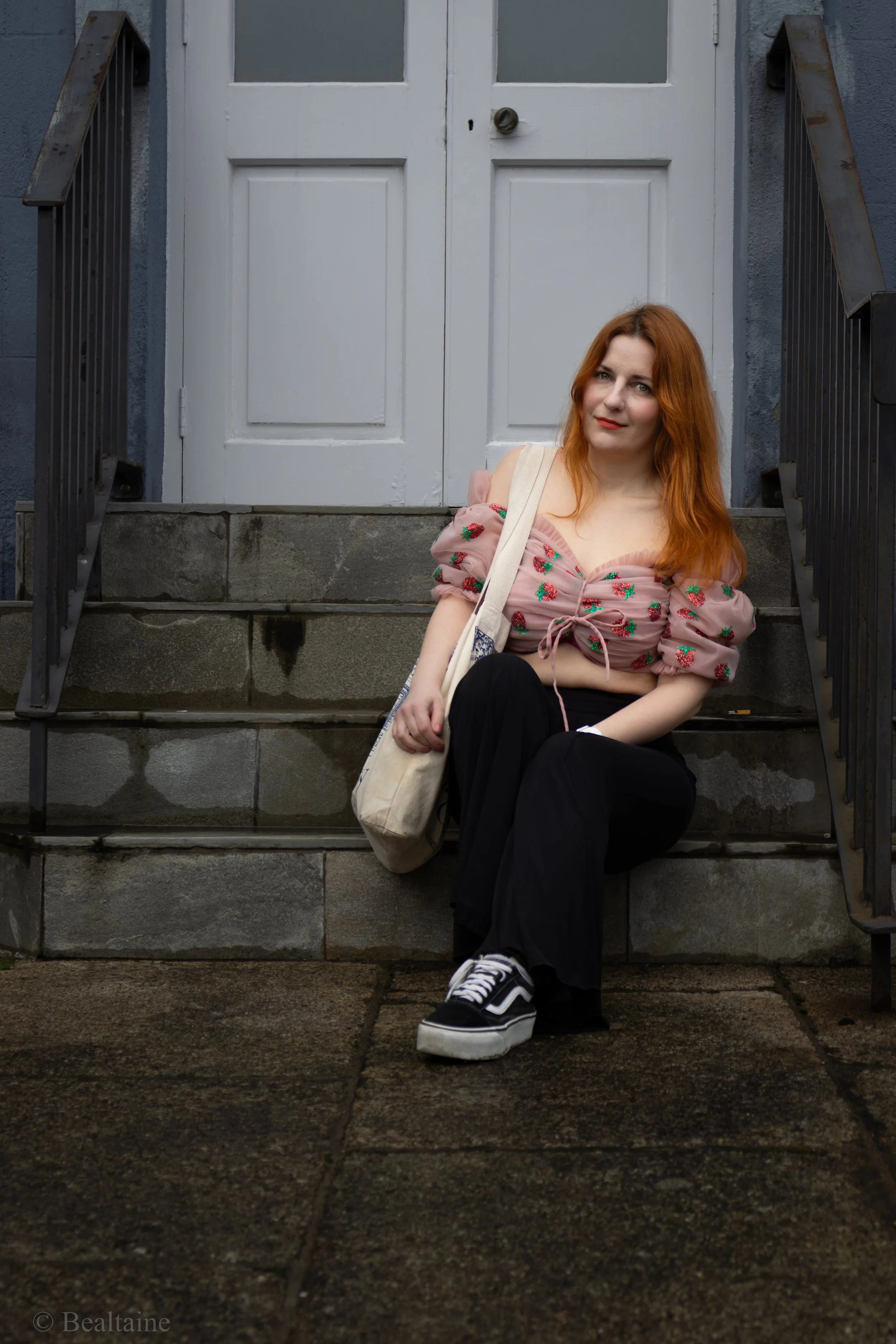 A young woman with red hair sitting on stone steps outside a building with white double doors. She is wearing a pink, puff-sleeve top with strawberry patterns, black pants, and black and white sneakers. She has a tote bag over her shoulder and is loo