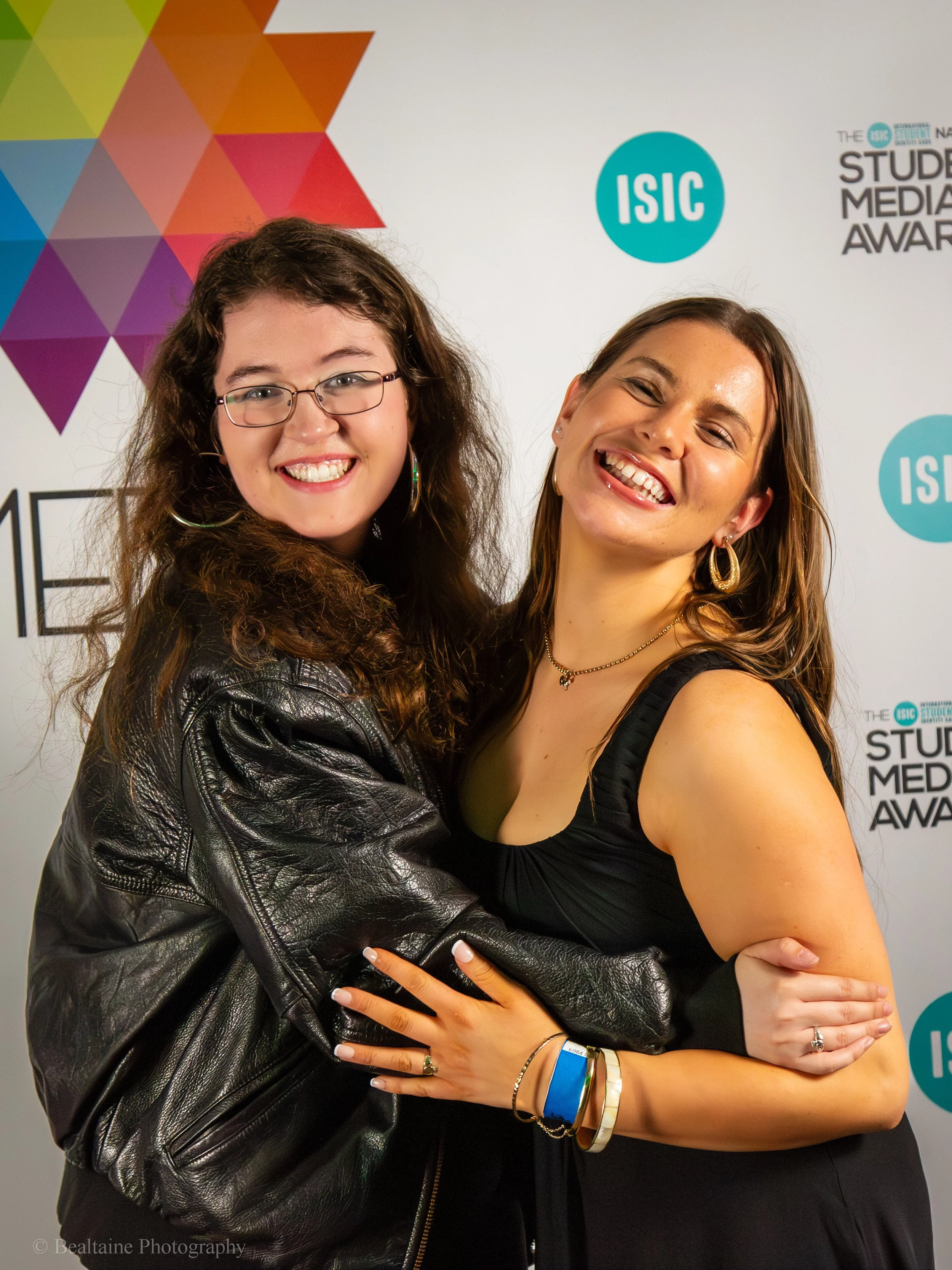 Two women smiling and hugging at the Student Media Awards event, with the ISC logo on the backdrop.
