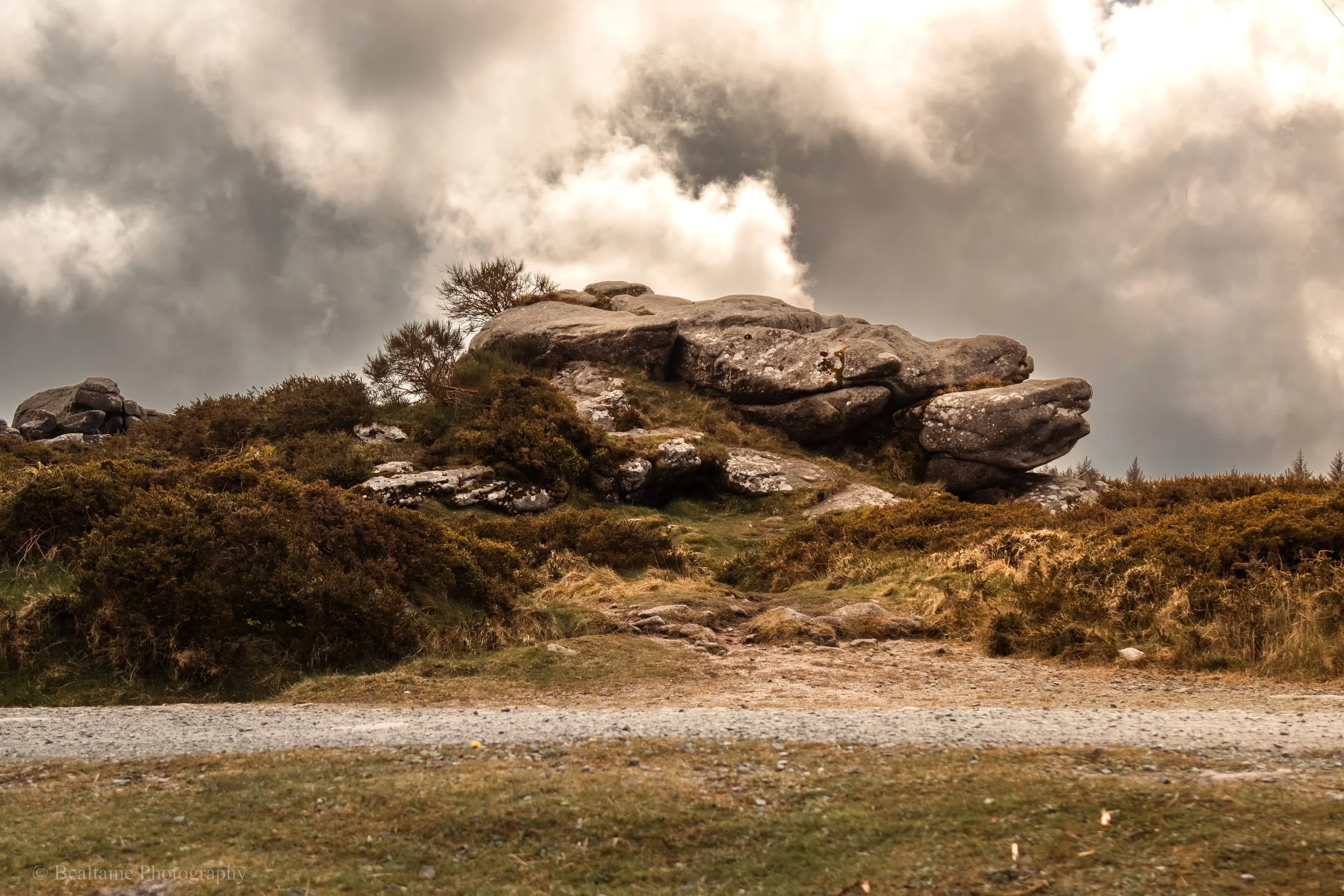 A rocky hillside with a dirt path in front, under a cloudy sky, with bushes and trees on the hill.