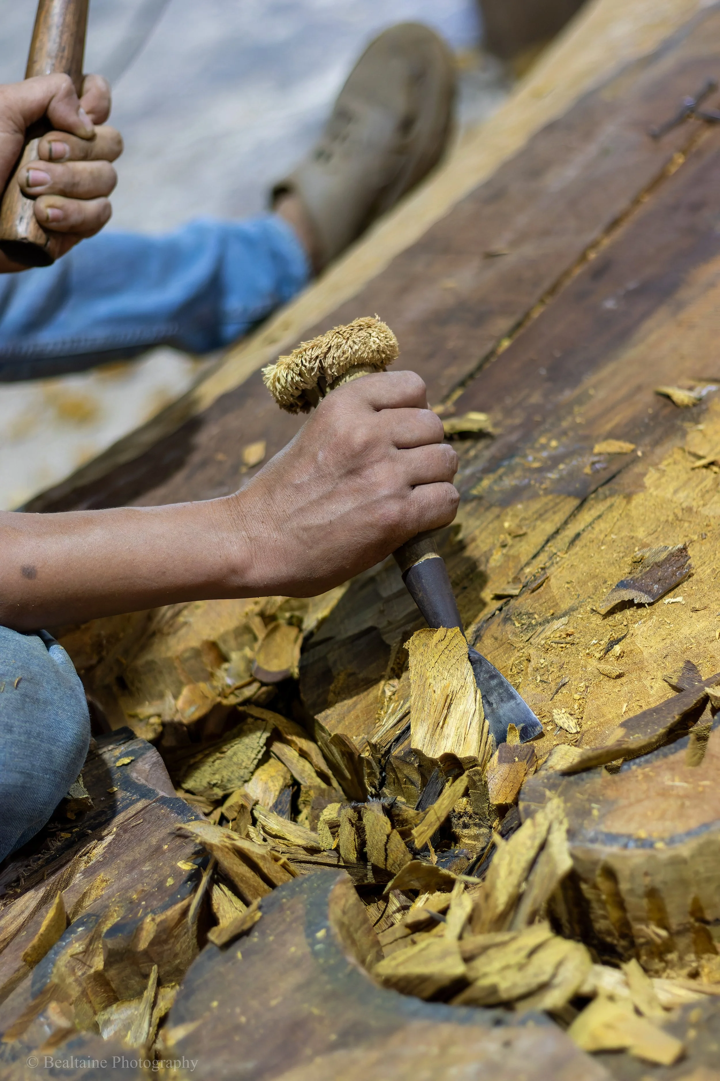 Close-up of a person's hand sawing a large piece of wood with a hand saw, surrounded by wood shavings.