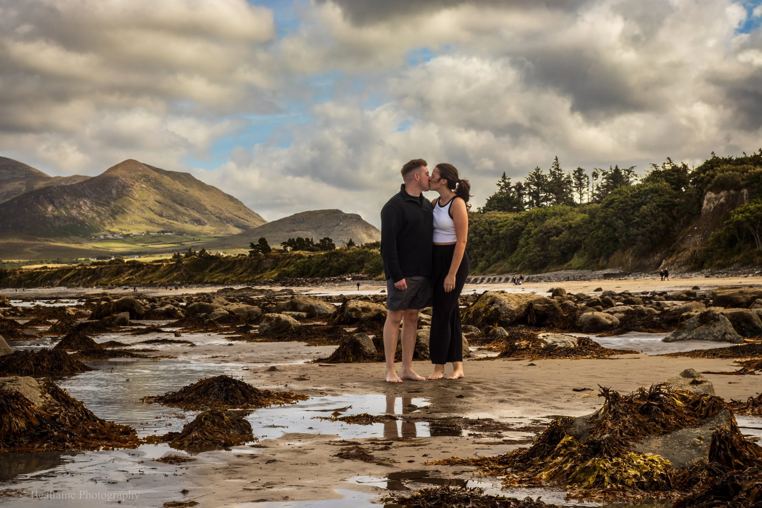 A couple kissing on a rocky beach with mountains and cloudy sky in the background.
