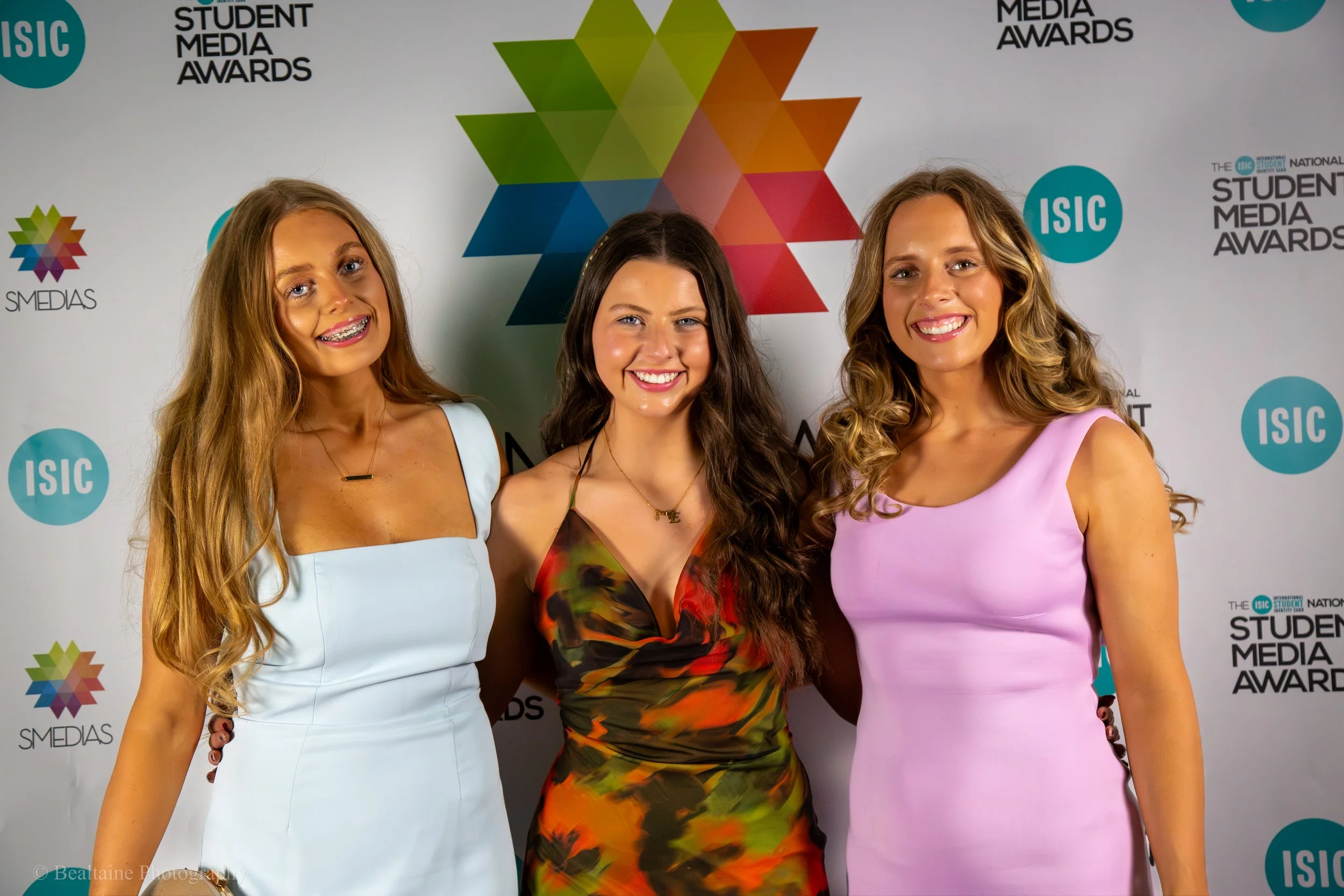 Three young women dressed in formal attire posing together at the Student Media Awards event with a colorful geometric backdrop and event logos.