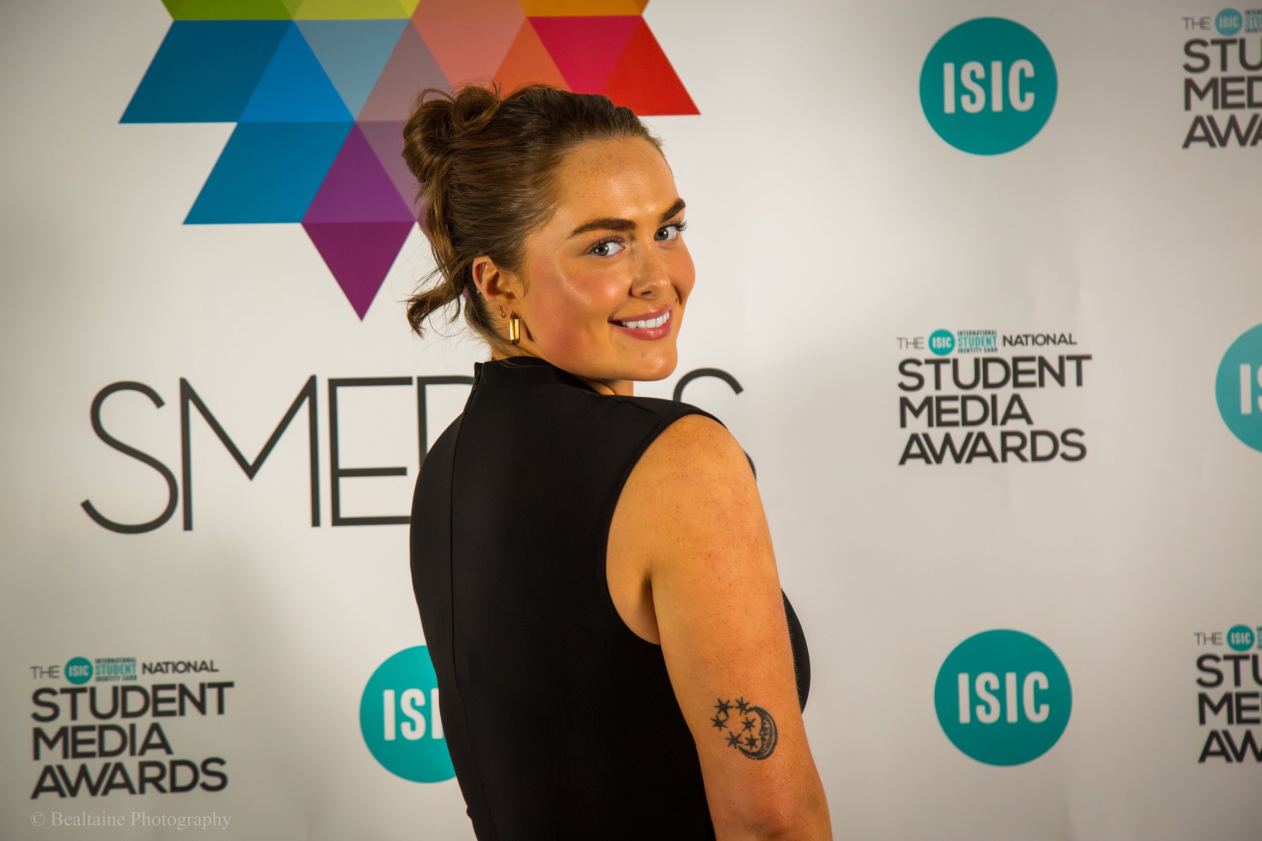 A smiling woman with brown hair tied in a bun, wearing a black sleeveless dress, standing in front of a backdrop with colorful geometric shapes and text that reads 'STUDENT MEDIA AWARDS' and 'ISC'.