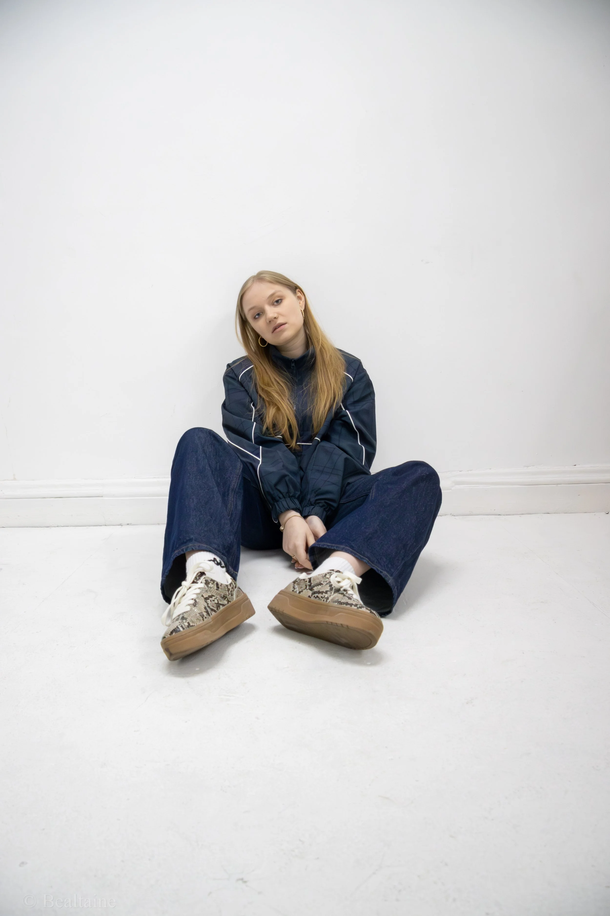 A young woman with long blonde hair, wearing a dark jacket and casual jeans, sitting on a white floor against a white wall, with her legs crossed and her head tilted, looking at the camera.