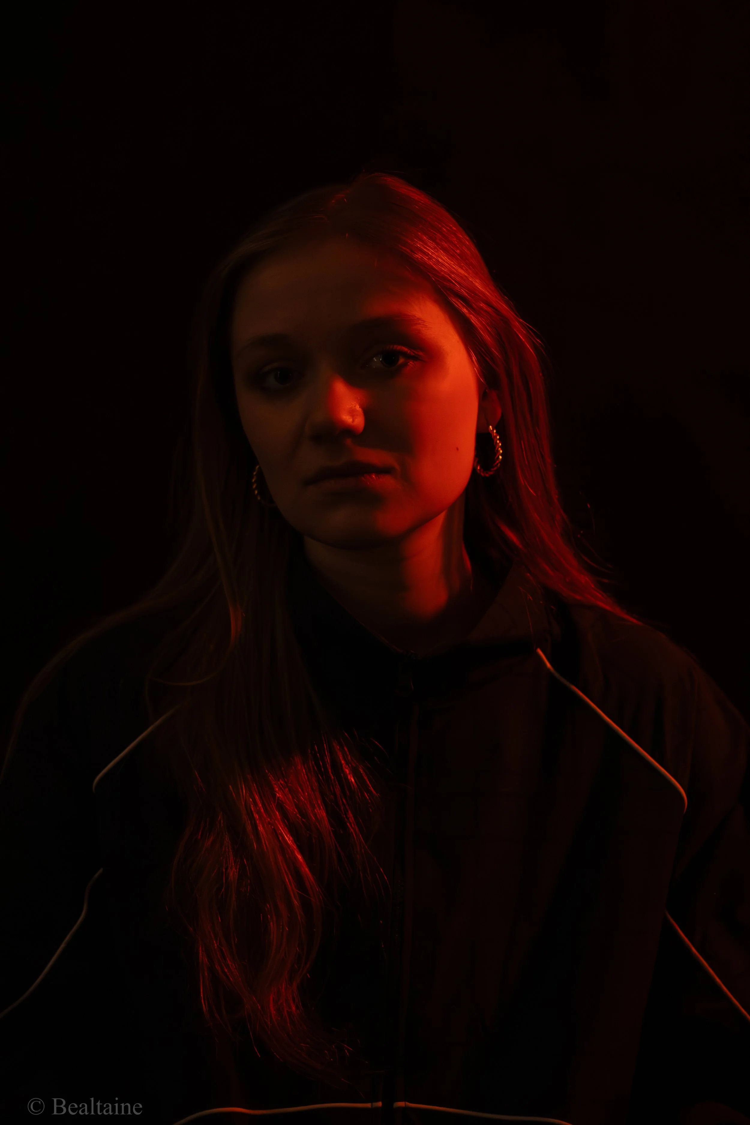 A young woman with long hair and hoop earrings illuminated by red and orange lighting, looking at the camera against a dark background.