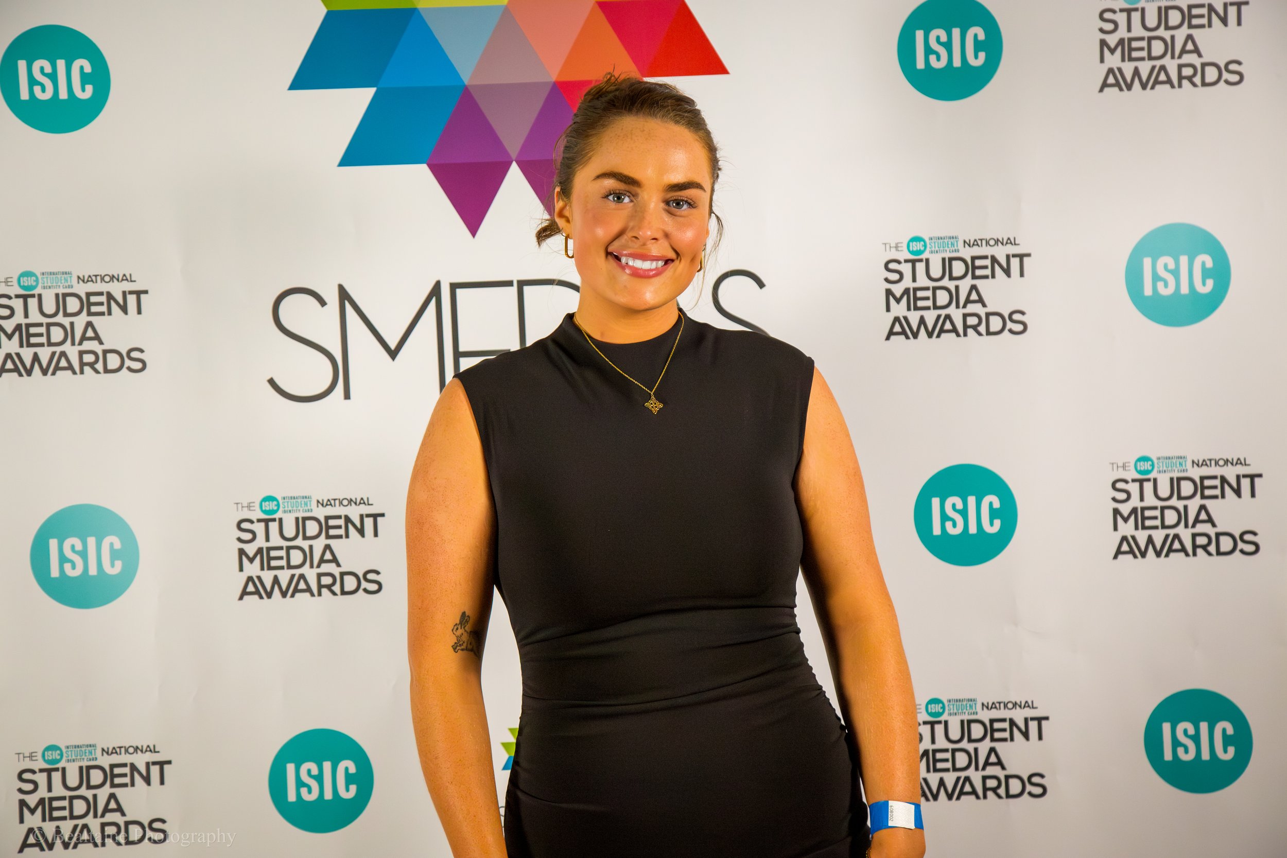A young woman with brown hair tied back, wearing a black sleeveless dress and gold necklace, smiling at the camera, standing in front of a backdrop with 'ISIC', 'Student Media Awards', and colorful geometric designs.
