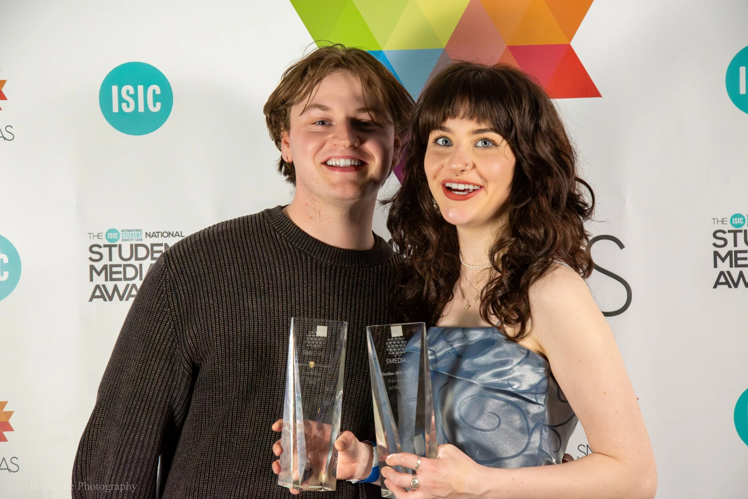 Two young adults, a man and a woman, smiling and holding awards or trophies at an awards ceremony. The background features the text 'ISIC' and 'STUDENT MEDIA AWARDS', with colorful geometric shapes.
