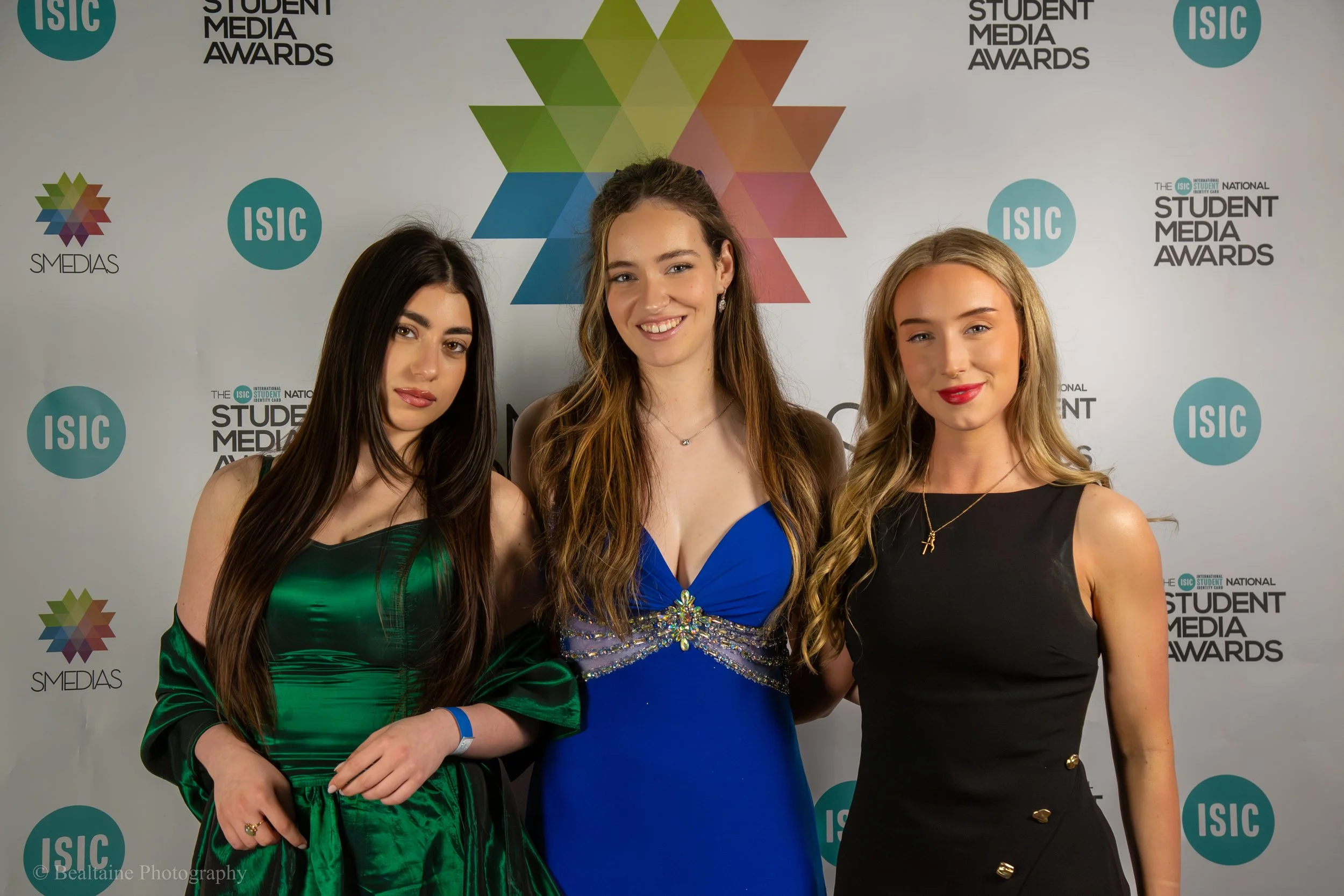 Three young women dressed in formal evening gowns standing together at the Student Media Awards event, with a backdrop displaying the event logos and colorful geometric design.